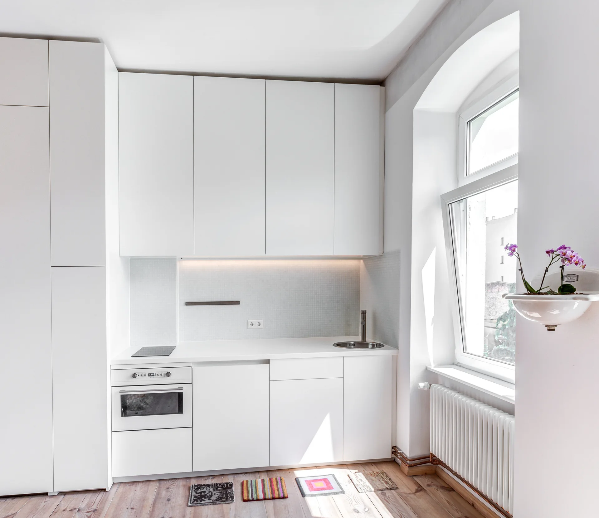 A compact minimalist kitchen featuring flat-front white cabinets, a small tiled backsplash, an integrated oven, and a large arched window.