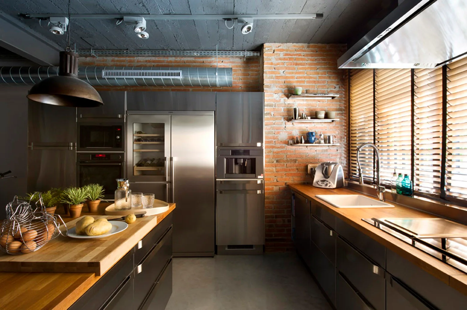 An industrial kitchen features an exposed brick wall, stainless steel appliances, dark grey lower cabinets with wood countertops, and a central island.