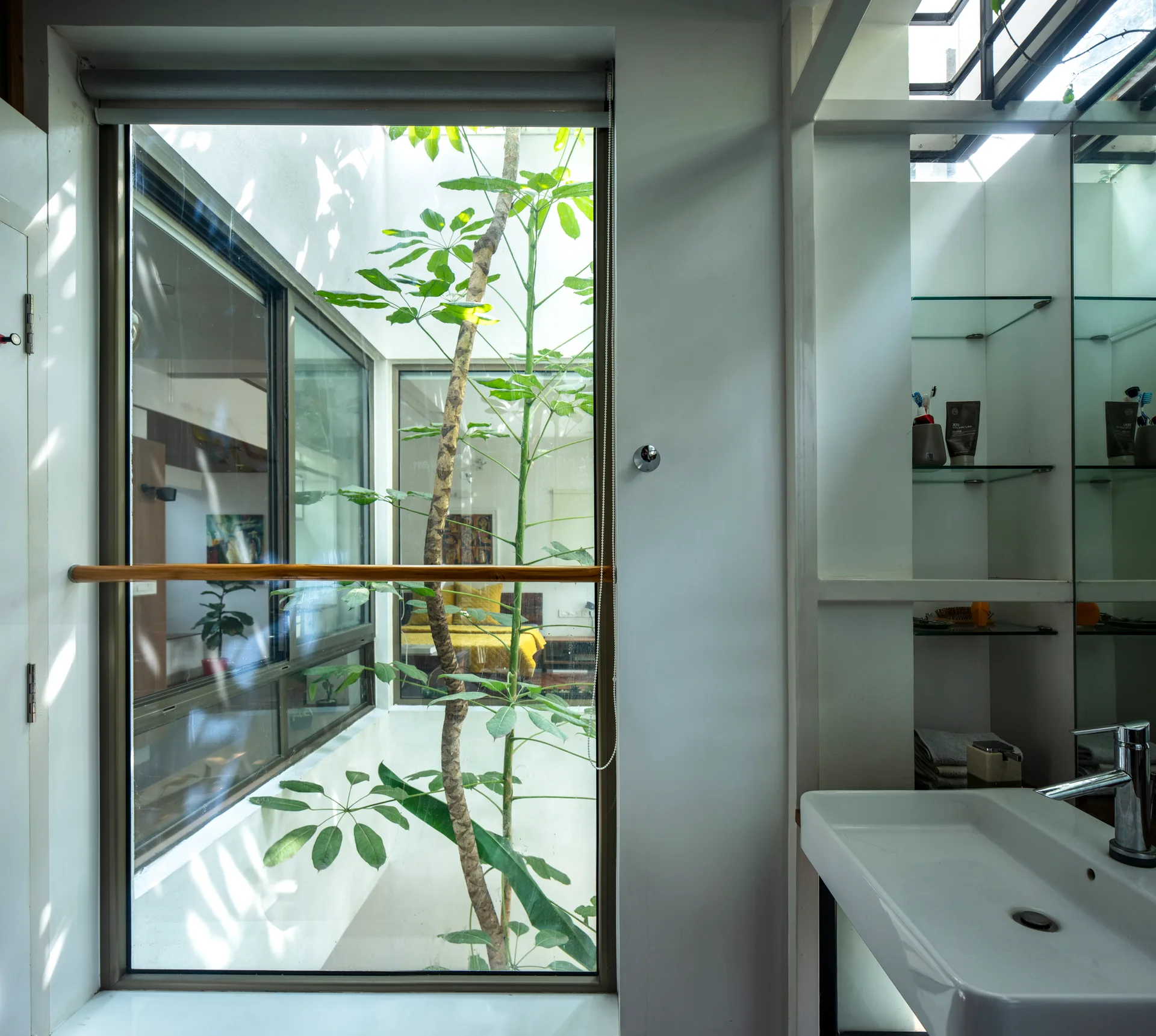 A modern white sink and glass-shelved alcove stand next to a large window overlooking a plant-filled interior lightwell.