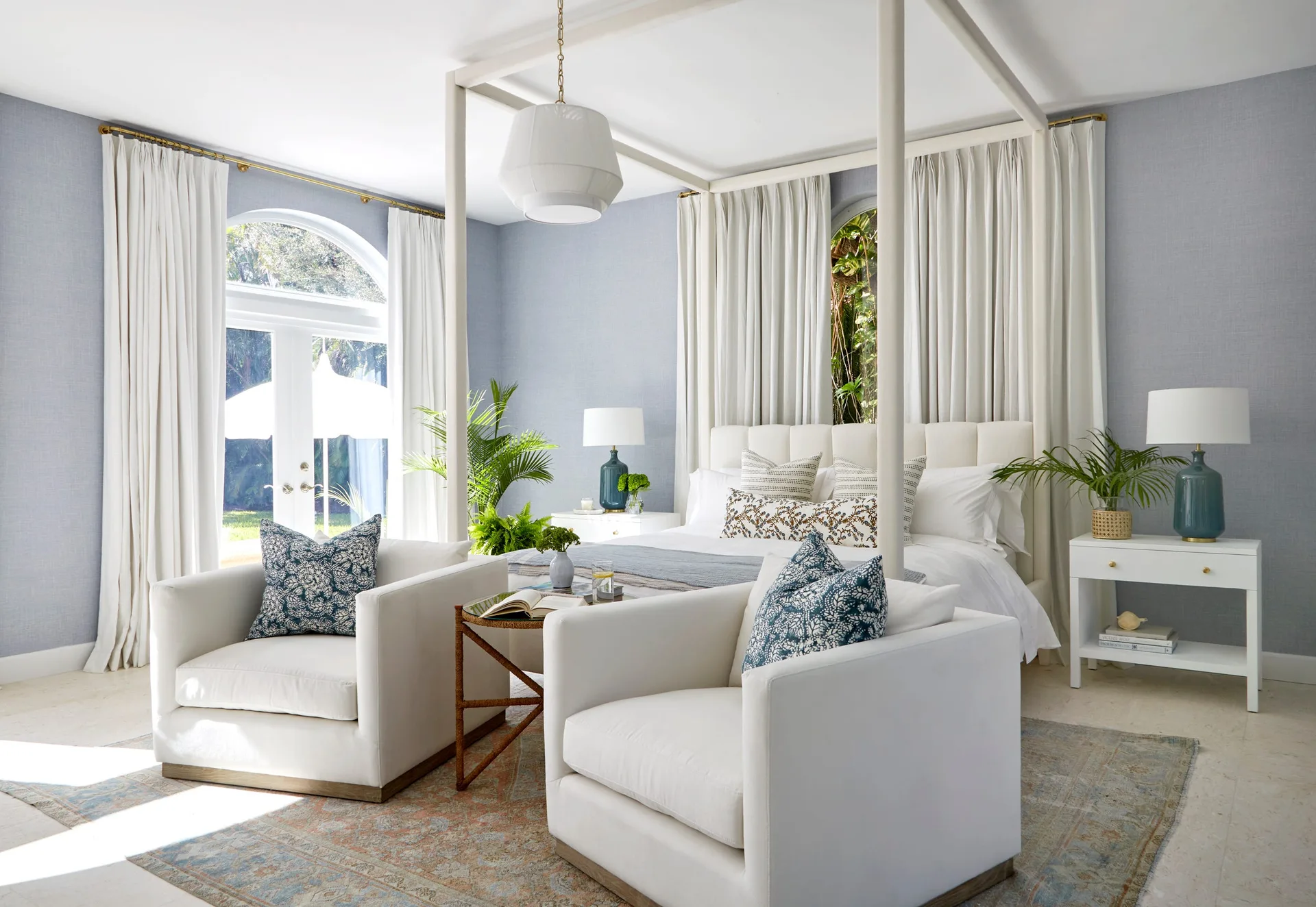 A light blue bedroom featuring a white canopy bed, white drapes, two white armchairs with blue patterned pillows, and blue table lamps.