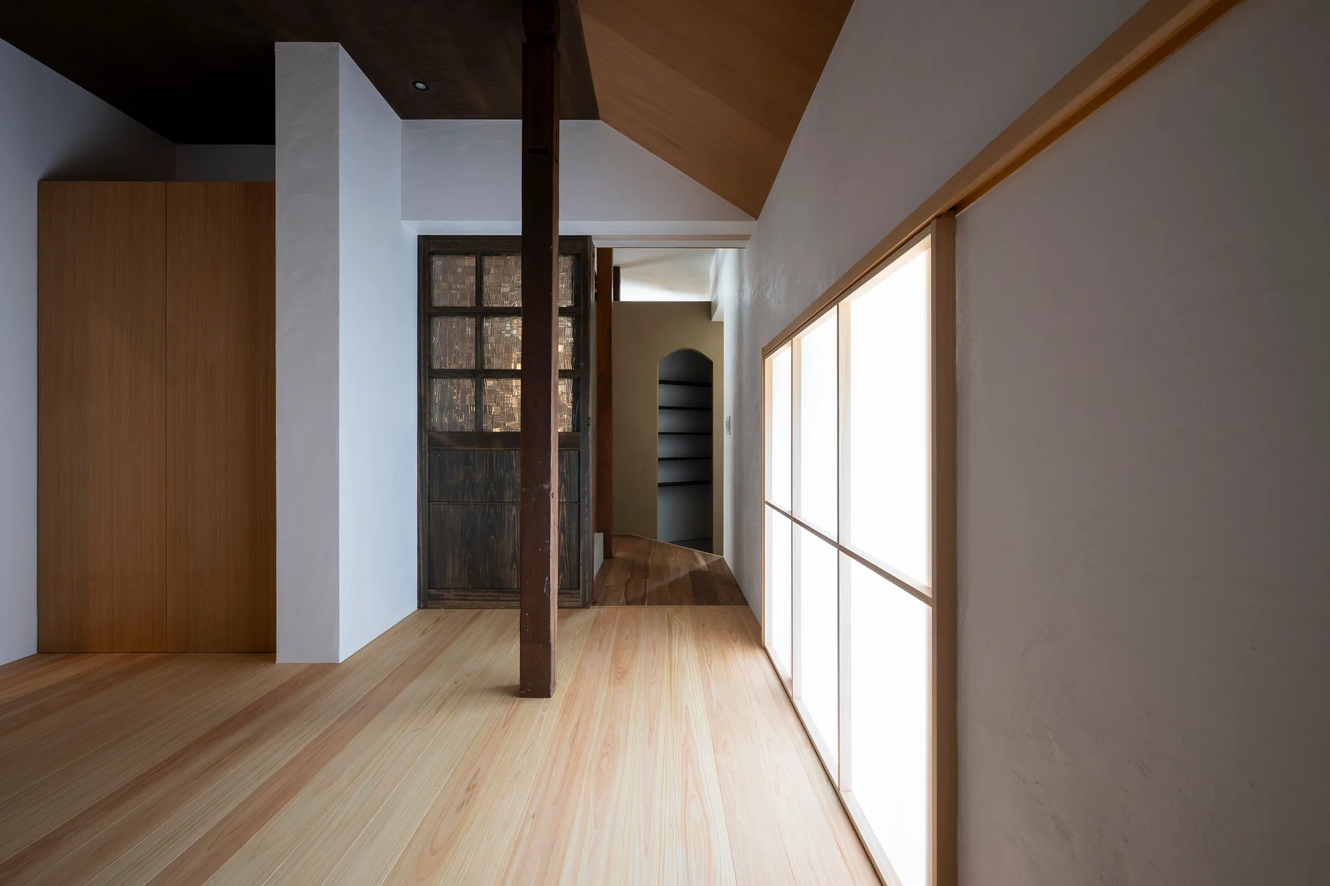 A minimalist hallway features light wood flooring, white plaster walls, a central timber post, and a large illuminated shoji-style screen window.