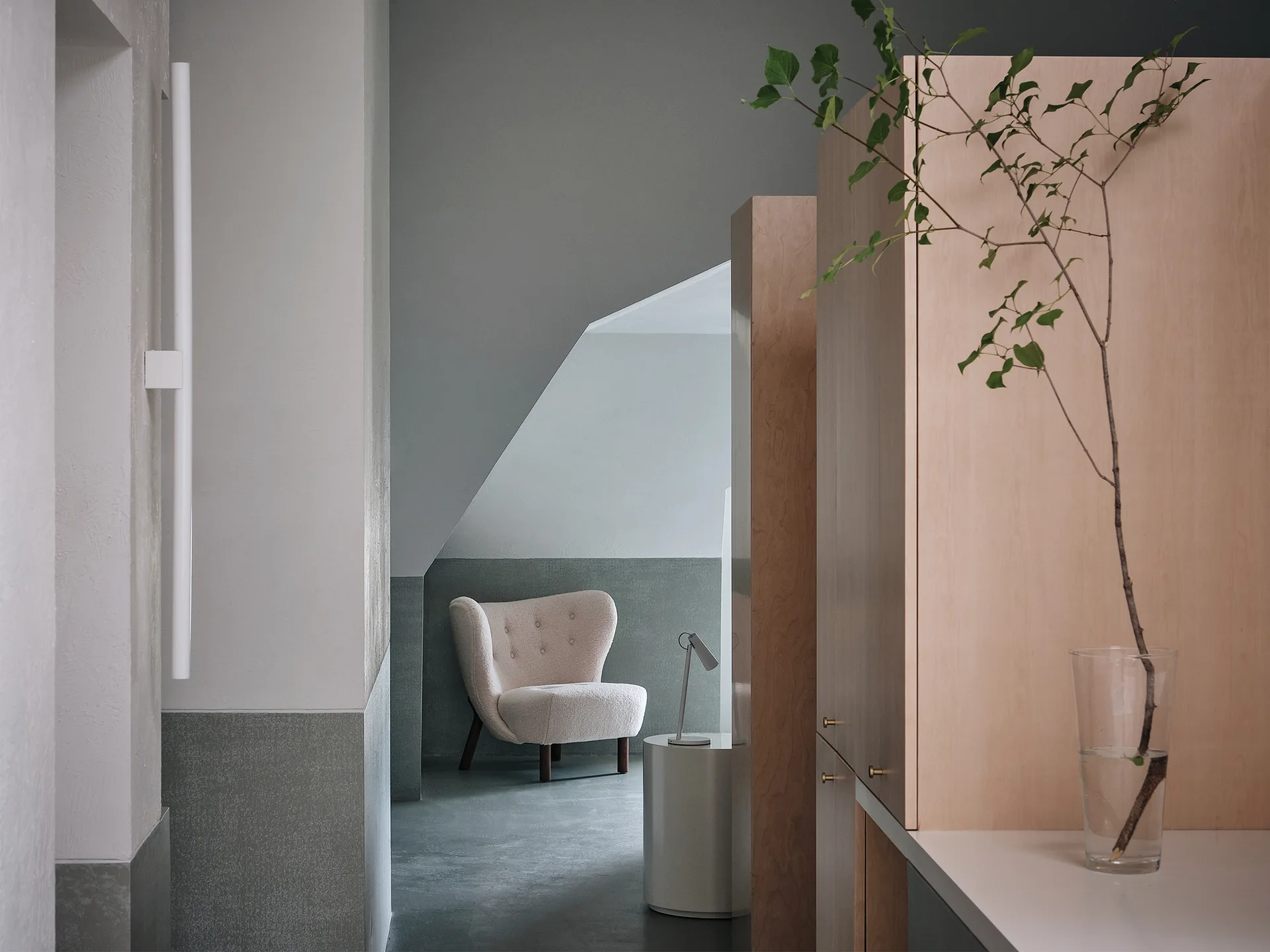 A minimalist interior featuring a cream tufted armchair, light wood cabinets, and two-tone painted walls with a cylindrical side table.