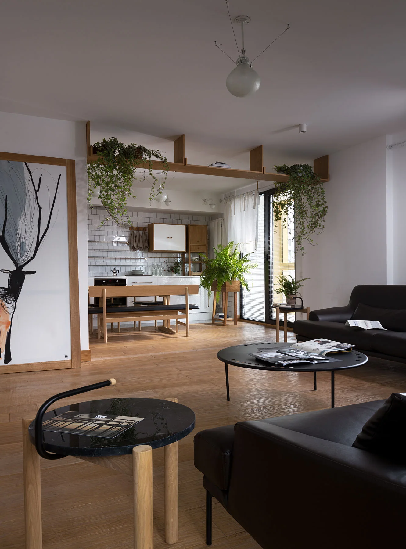 An open-concept living room and kitchen featuring black leather sofas, a wooden dining table, and a high-mounted wooden shelf with trailing plants.