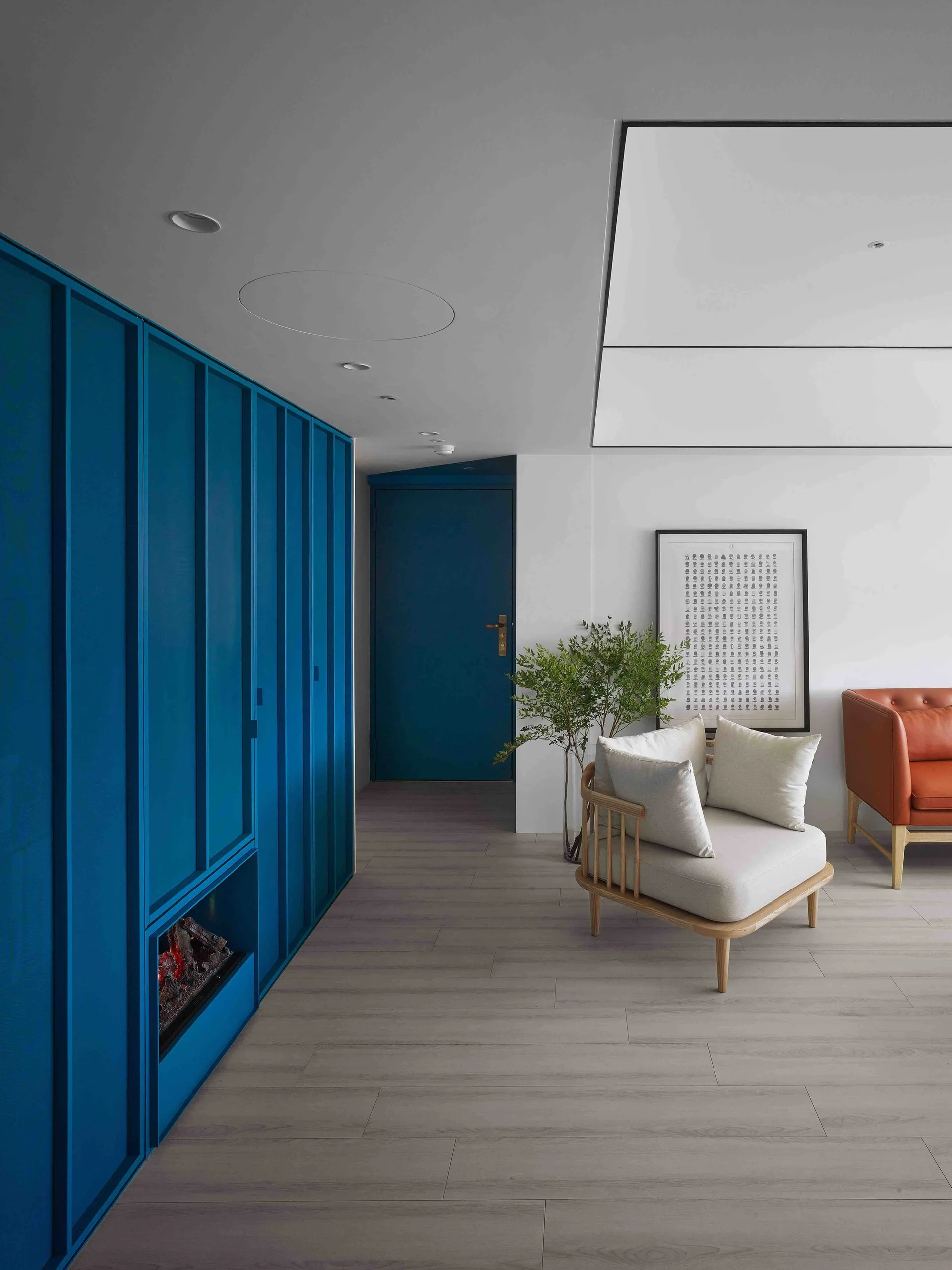 A contemporary living room featuring a solid blue wall of built-in cabinets with an integrated fireplace, a light wood armchair, and wood-plank flooring.