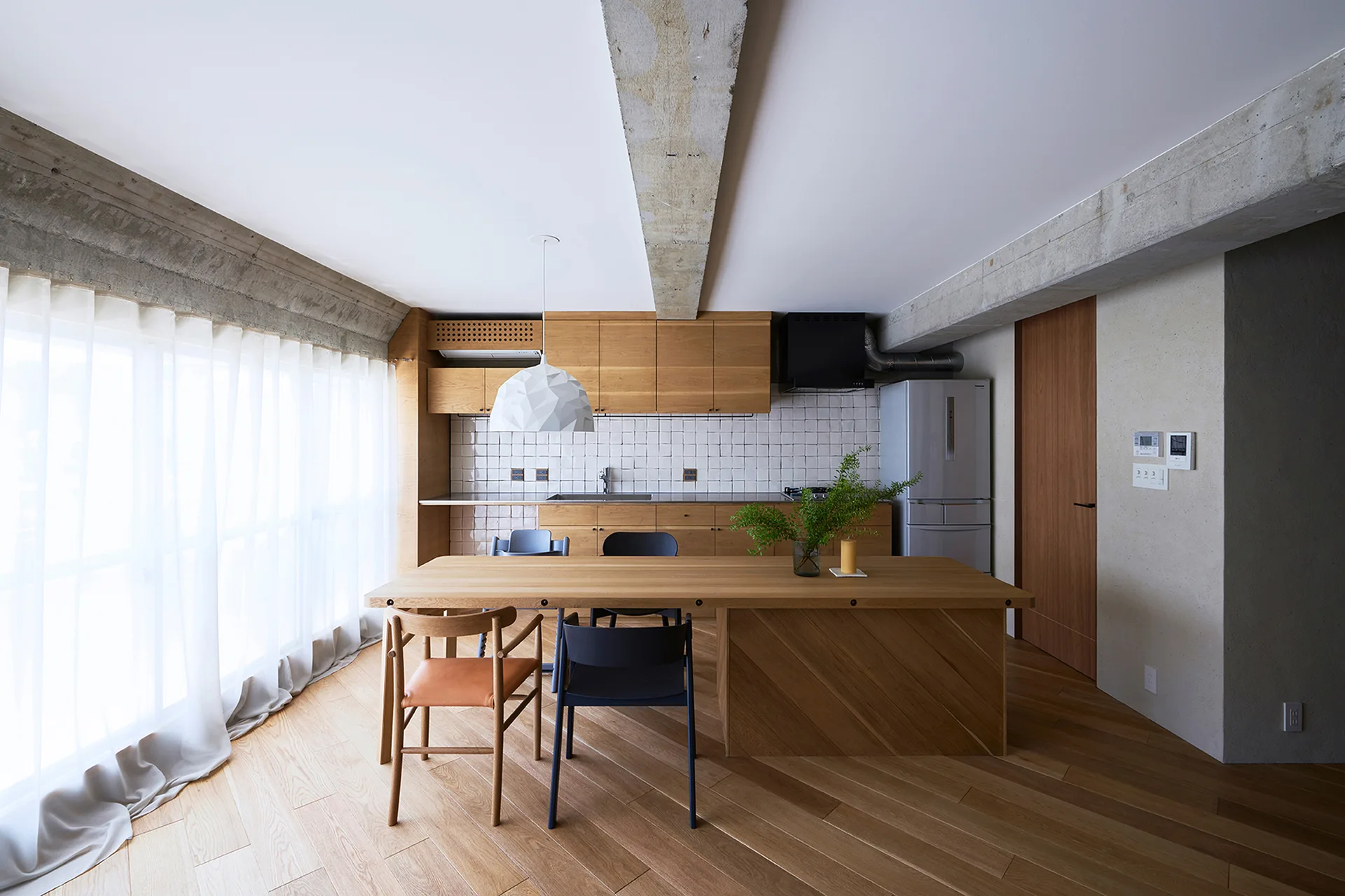 A modern kitchen features light wood cabinets, an oak dining table, a white tiled backsplash, and exposed raw concrete beams on the ceiling.