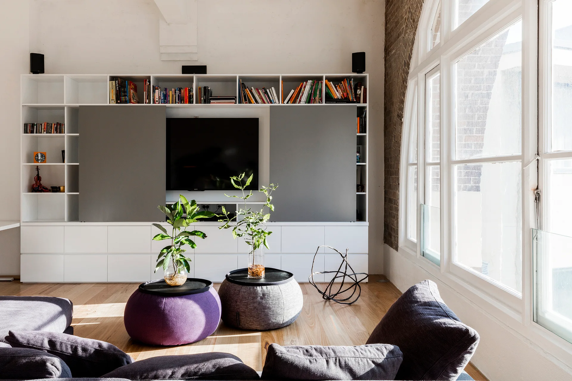 A bright living room features a white built-in wall unit with sliding grey panels, two round poufs with plants, and a large arched window.