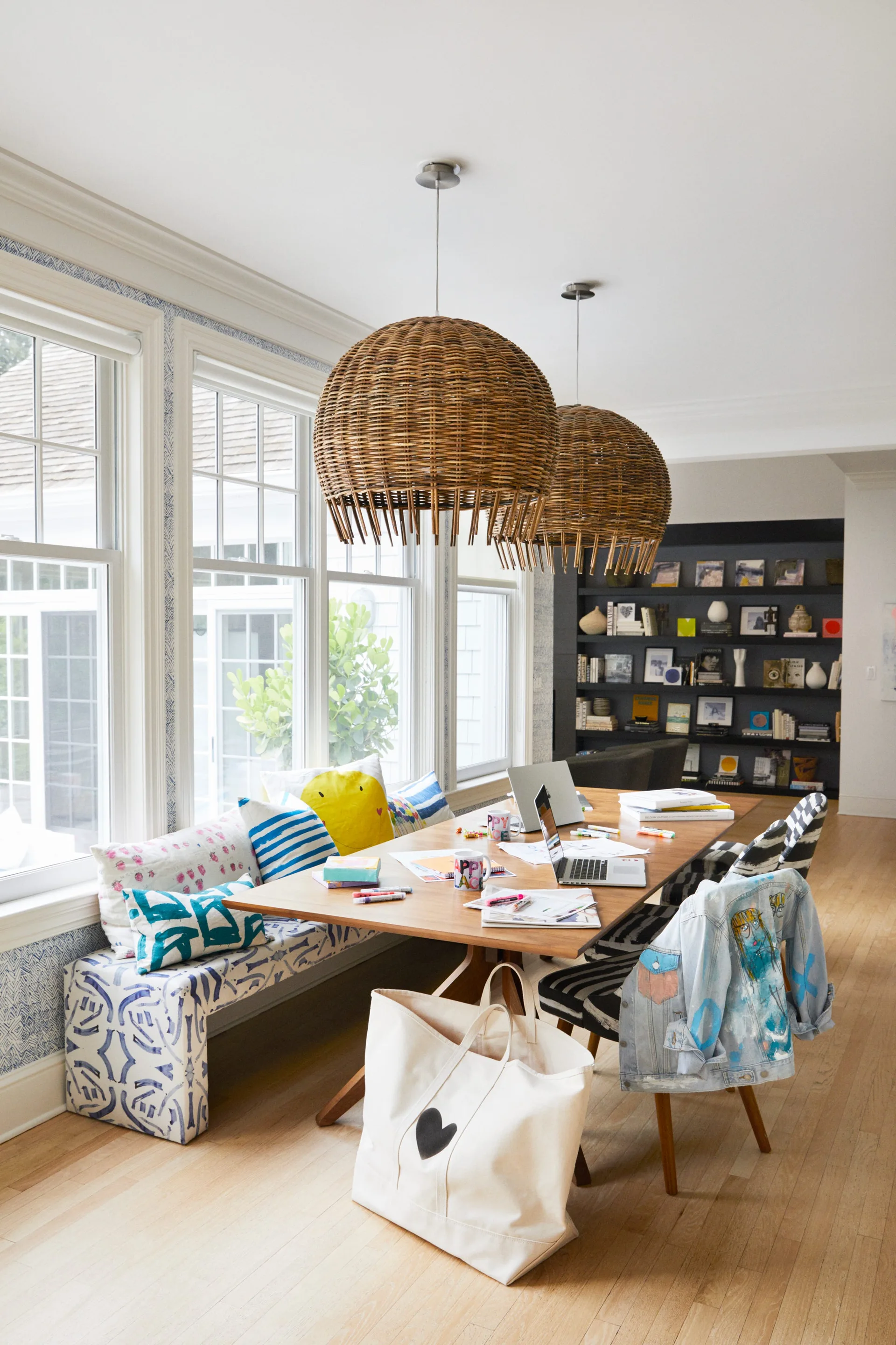 A wooden dining table features patterned banquette seating, striped chairs, and two large woven rattan pendant lights under a white ceiling.
