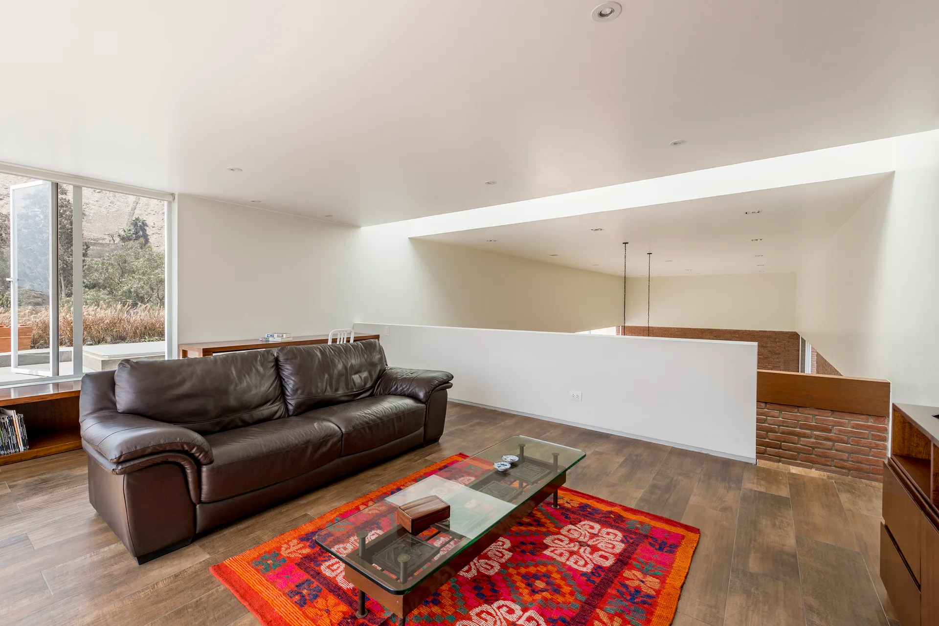 A bright loft living room features a dark brown leather sofa, a glass-top coffee table on a vibrant red patterned rug, and a large overhead skylight.
