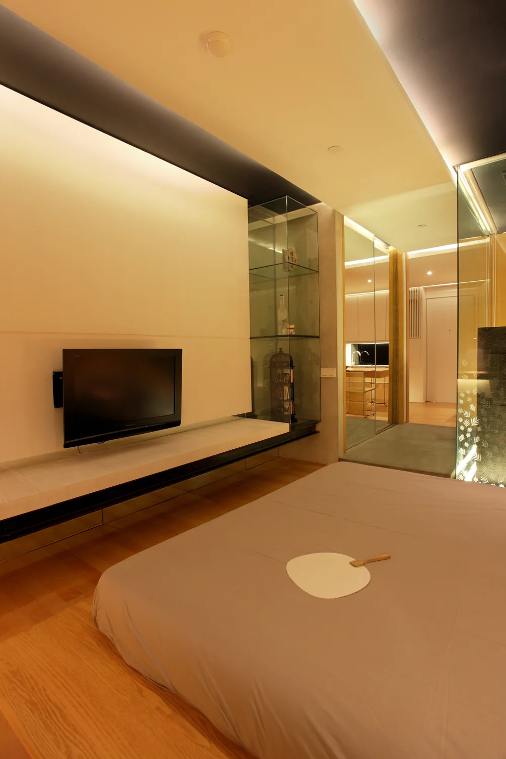 A minimalist bedroom features a low bed, a wall-mounted TV panel with a floating white console, and built-in glass display shelves.