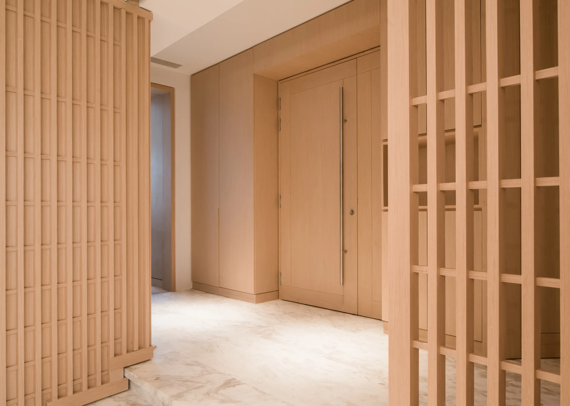 A modern hallway features light wood slatted screens, matching wall paneling with an integrated door, and polished white marble flooring.