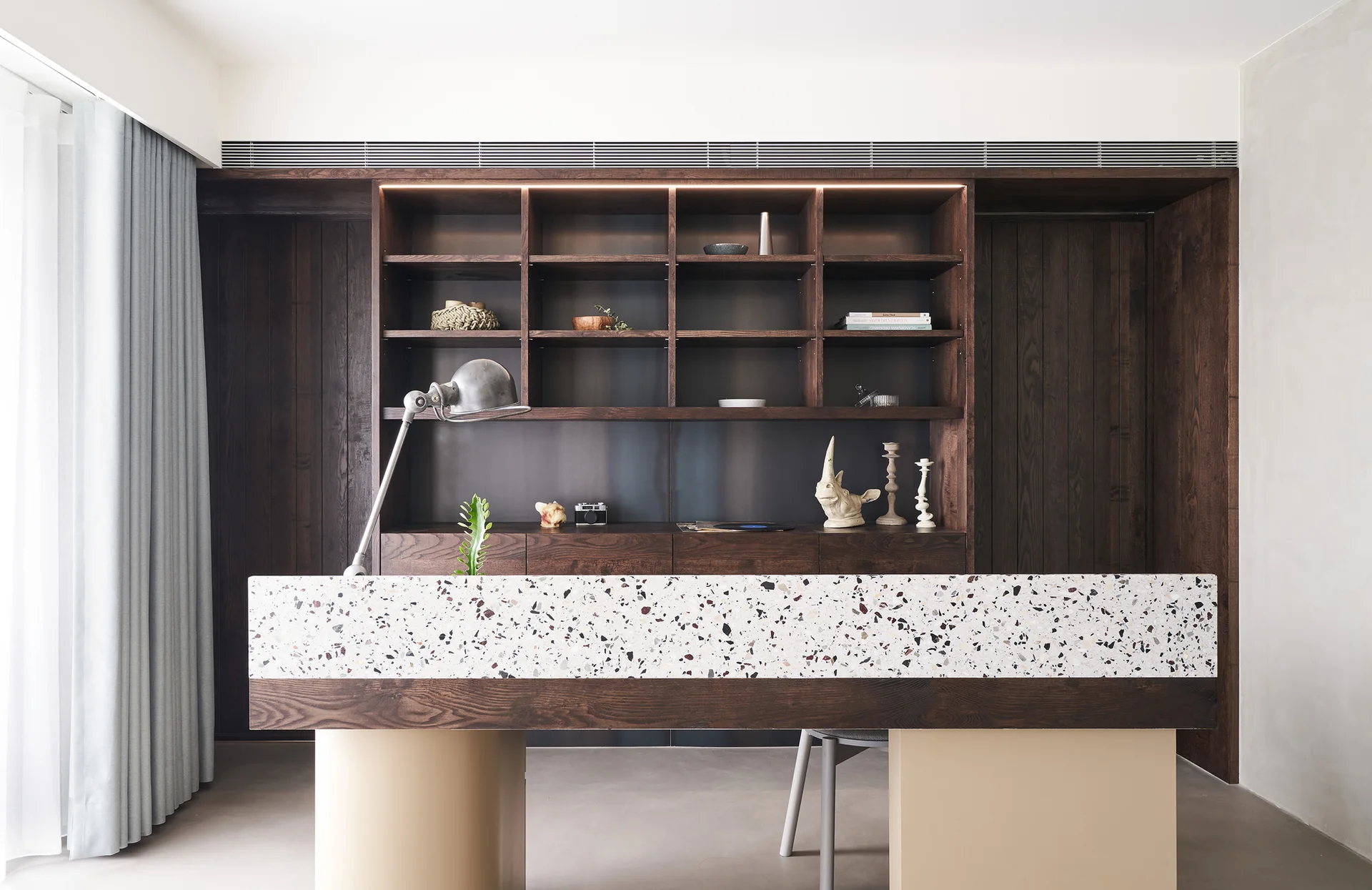 A home office featuring a dark wood built-in shelving unit behind a long desk with a speckled terrazzo countertop and task lamp.