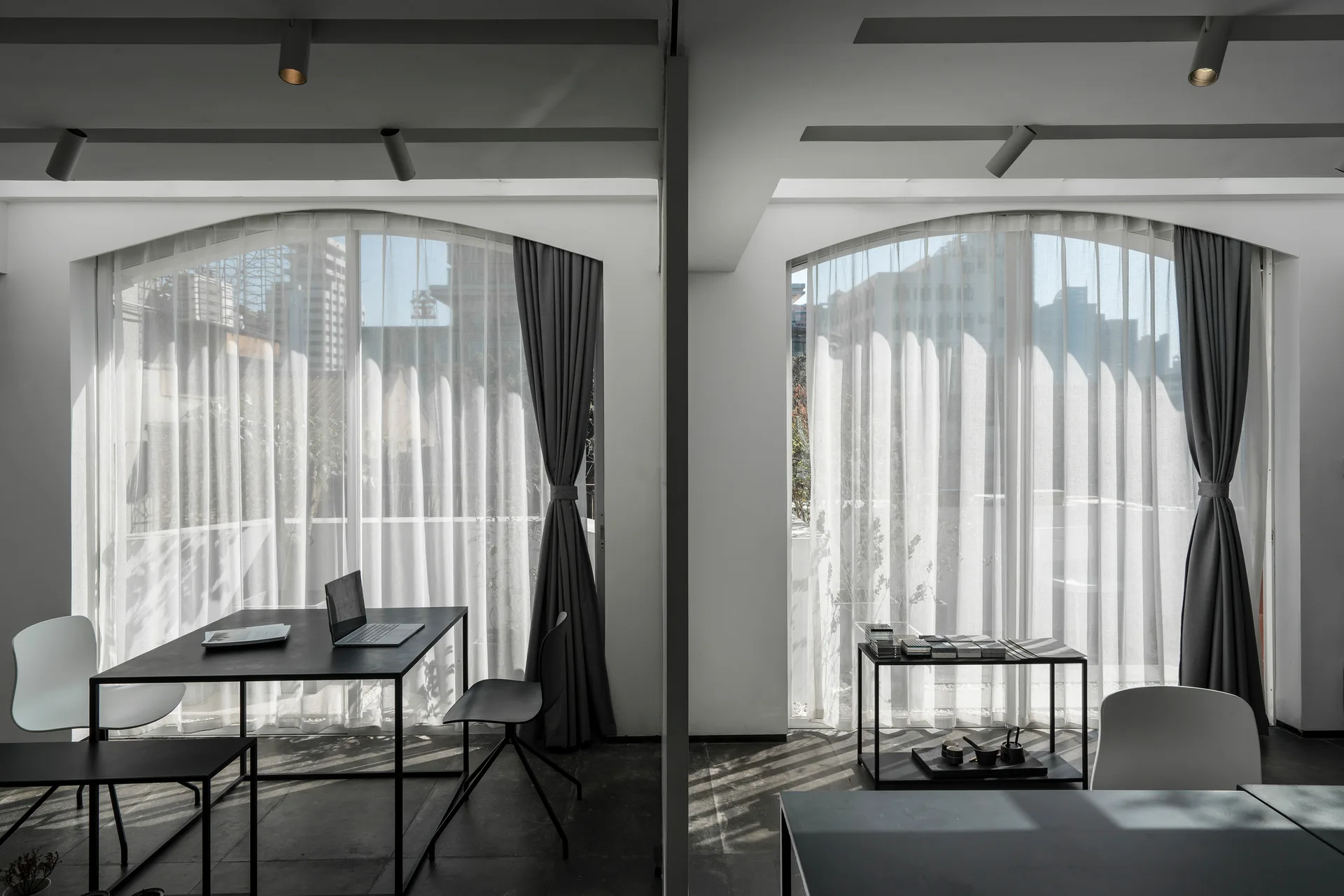 A minimalist black metal desk sits in front of a large arched window dressed with sheer white curtains and tied-back grey drapes in a modern workspace.