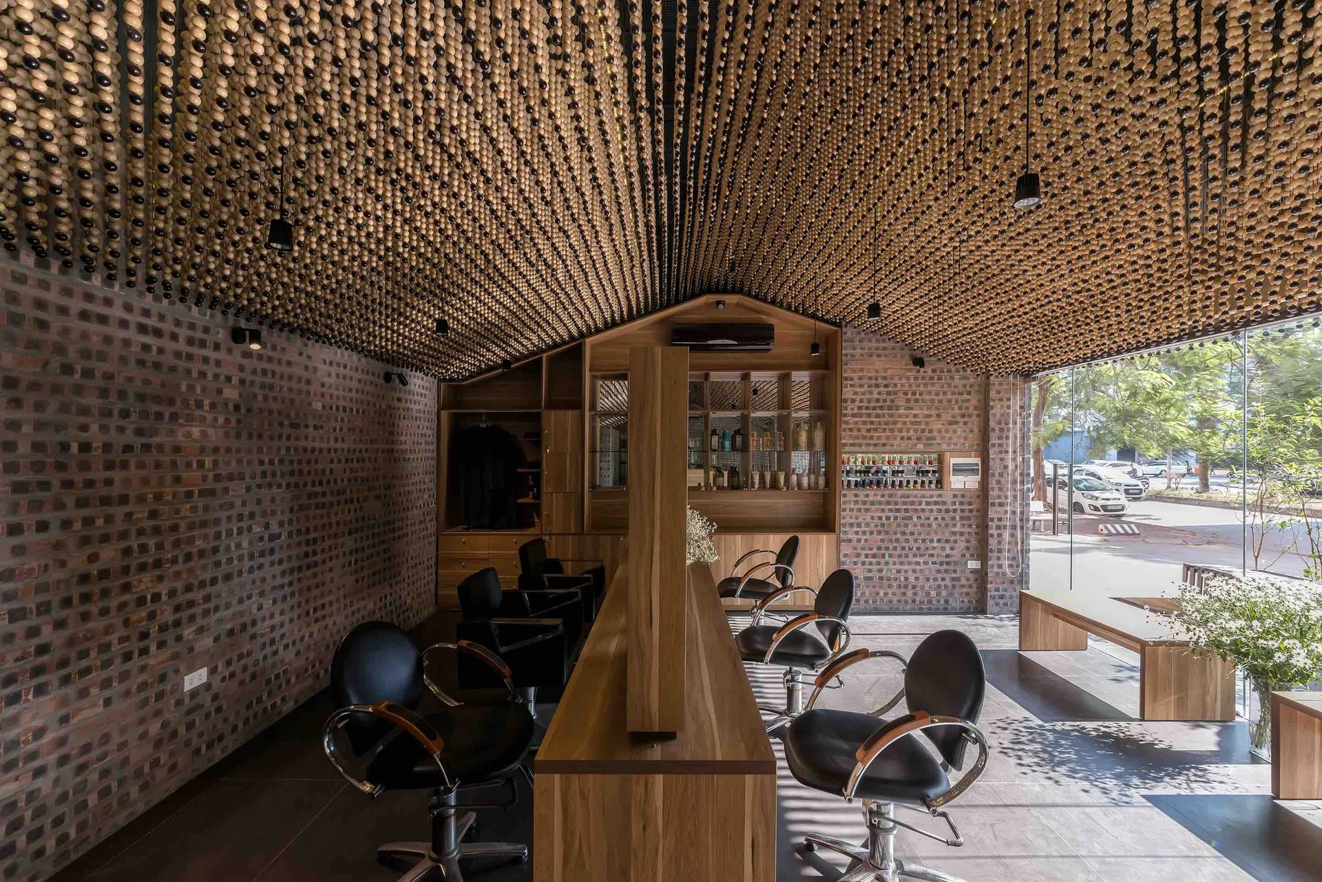 A modern salon interior featuring black styling chairs around a central wooden station beneath a dense ceiling installation of hanging wooden beads.