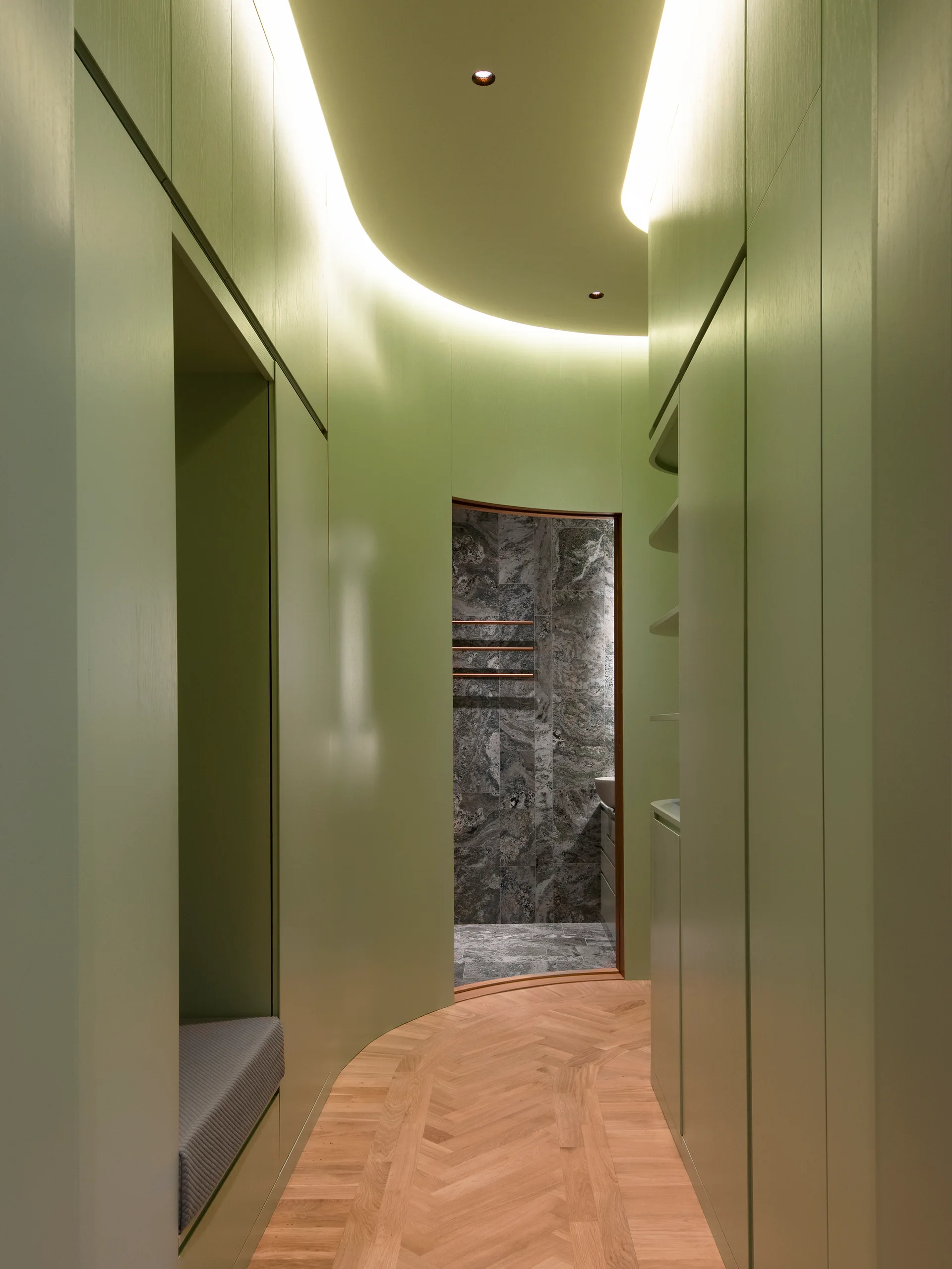 A curved sage green hallway featuring integrated custom cabinetry, light wood herringbone flooring, and cove lighting leading to a stone-tiled bathroom.