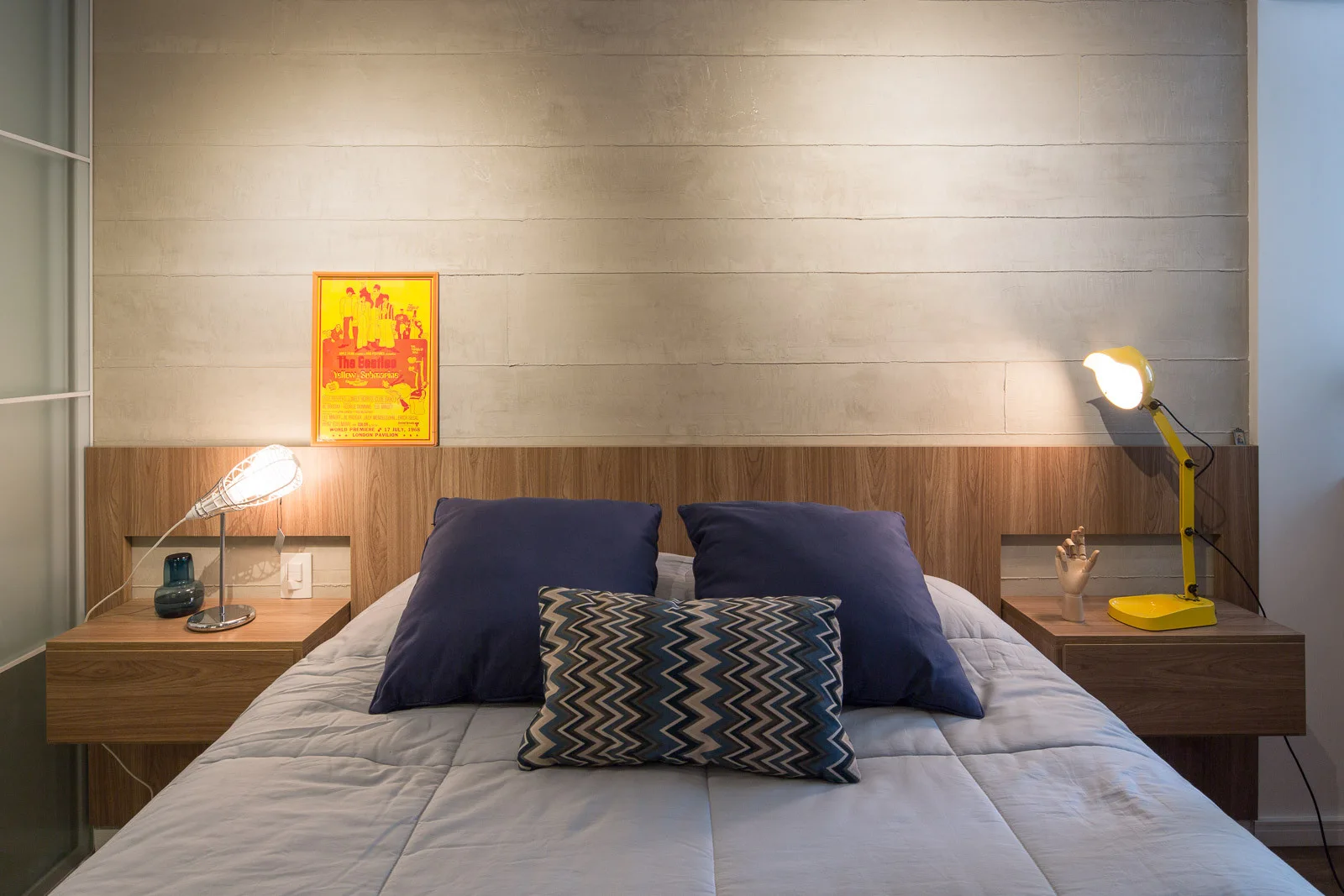A modern bedroom features a bed with blue pillows, a wood-toned headboard with floating nightstands, and two different glowing table lamps.