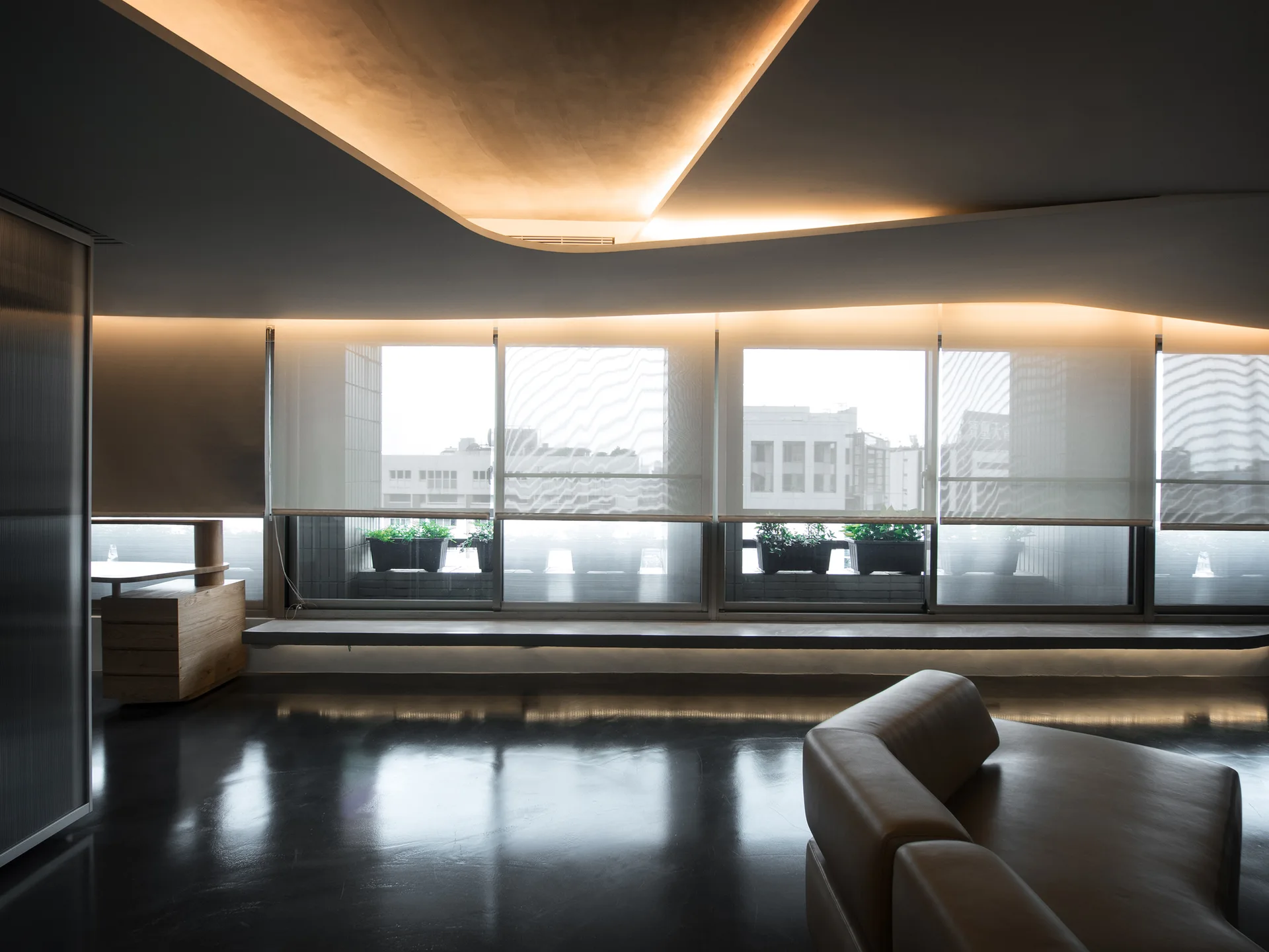 A modern living room features a curved leather sofa, polished dark floors, and large windows beneath an architectural ceiling with recessed cove lighting.