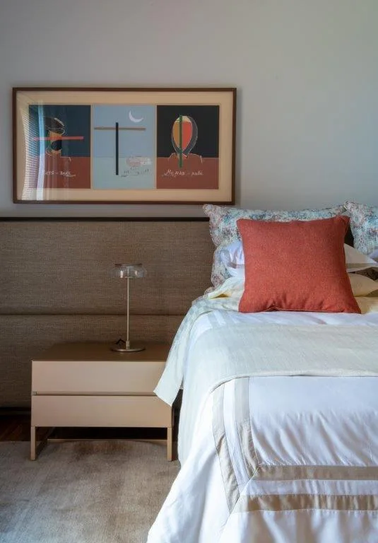 A bedroom features a bed with white linens and an orange pillow next to a beige nightstand, with a framed horizontal painting hanging above the headboard.