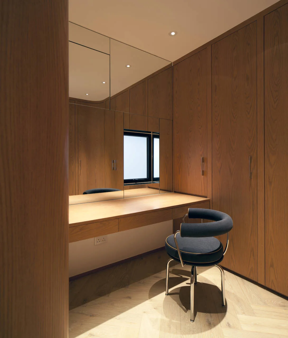A dressing room with wood paneled closets, an integrated oak vanity, a large wall mirror, and a circular chair.