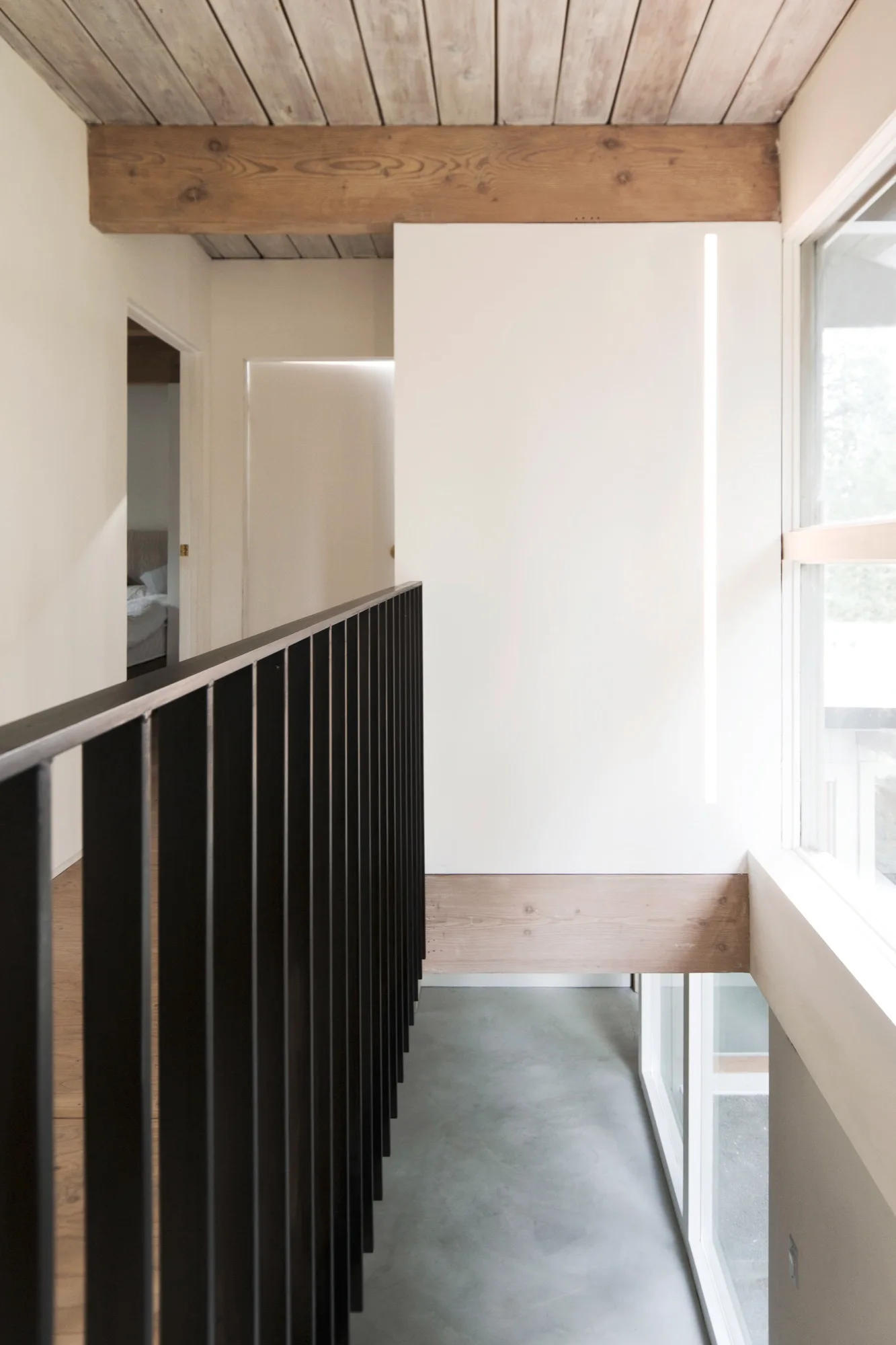 A modern residential landing featuring a black metal vertical railing, exposed wood ceiling beams, and clean white walls beside a large window.