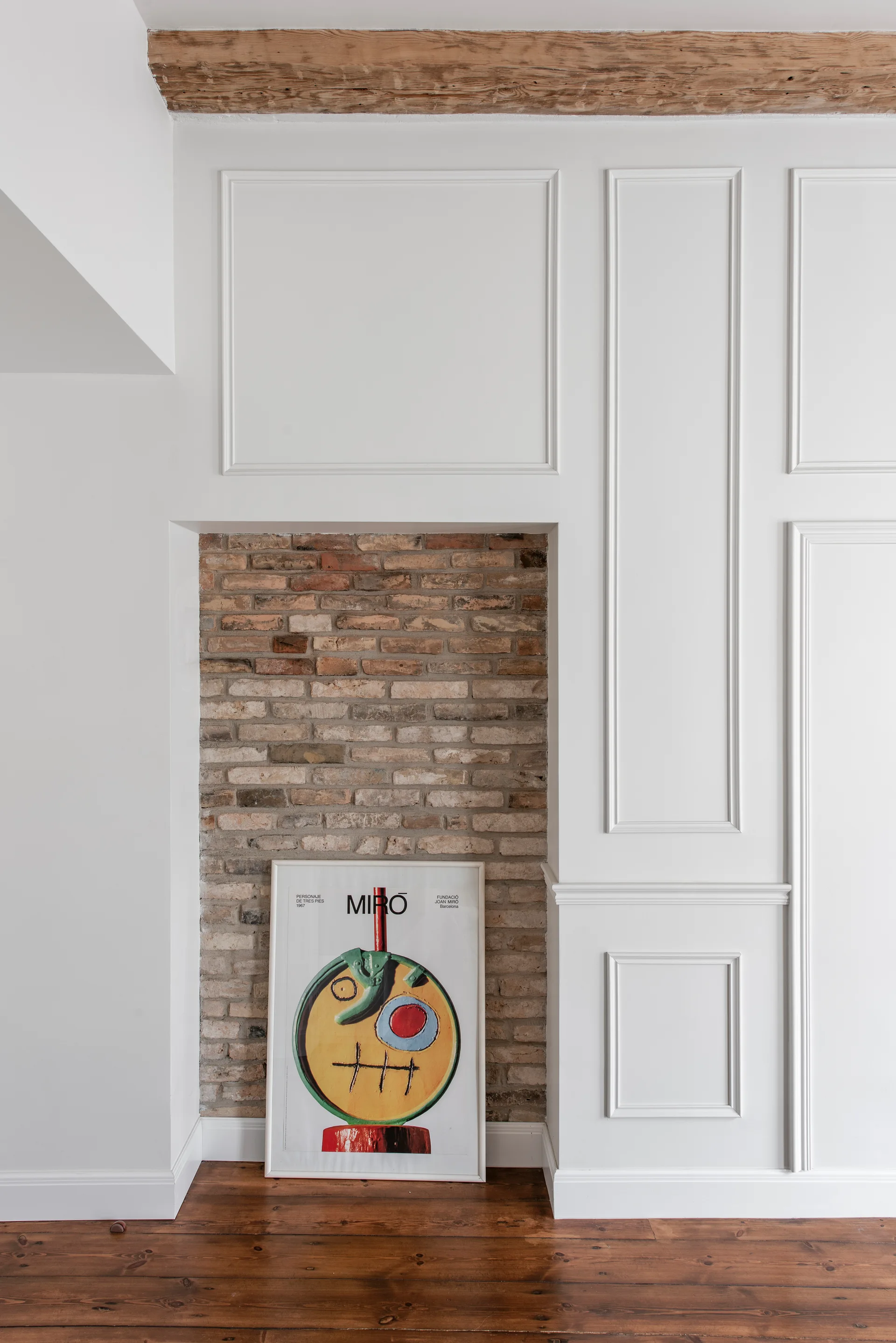 A framed art print rests on a wooden floor inside an exposed brick wall niche surrounded by white decorative paneling.
