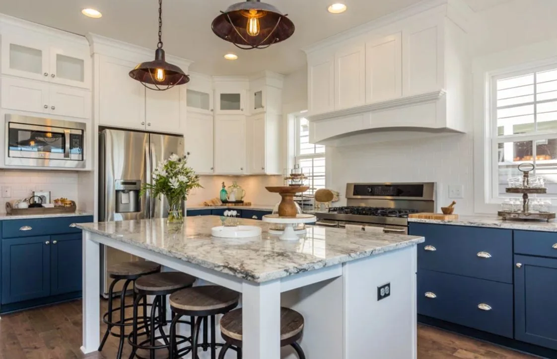 A kitchen features navy blue lower cabinets, white upper cabinets, a stone-topped island with wood-seated stools, and industrial pendant lights.