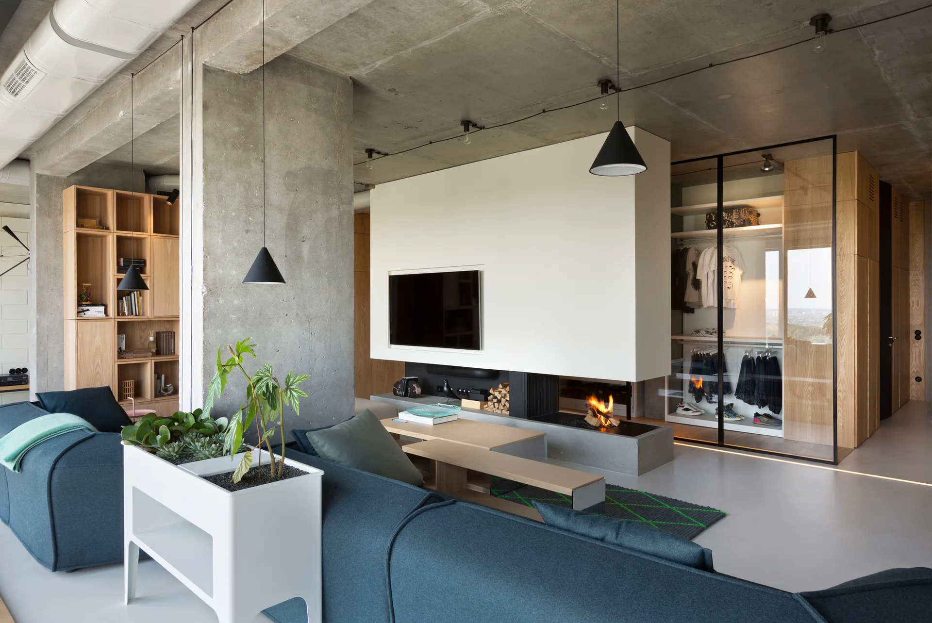 A modern industrial living room features a blue sofa, a central white fireplace partition, exposed concrete ceilings, and a glass-front wardrobe.