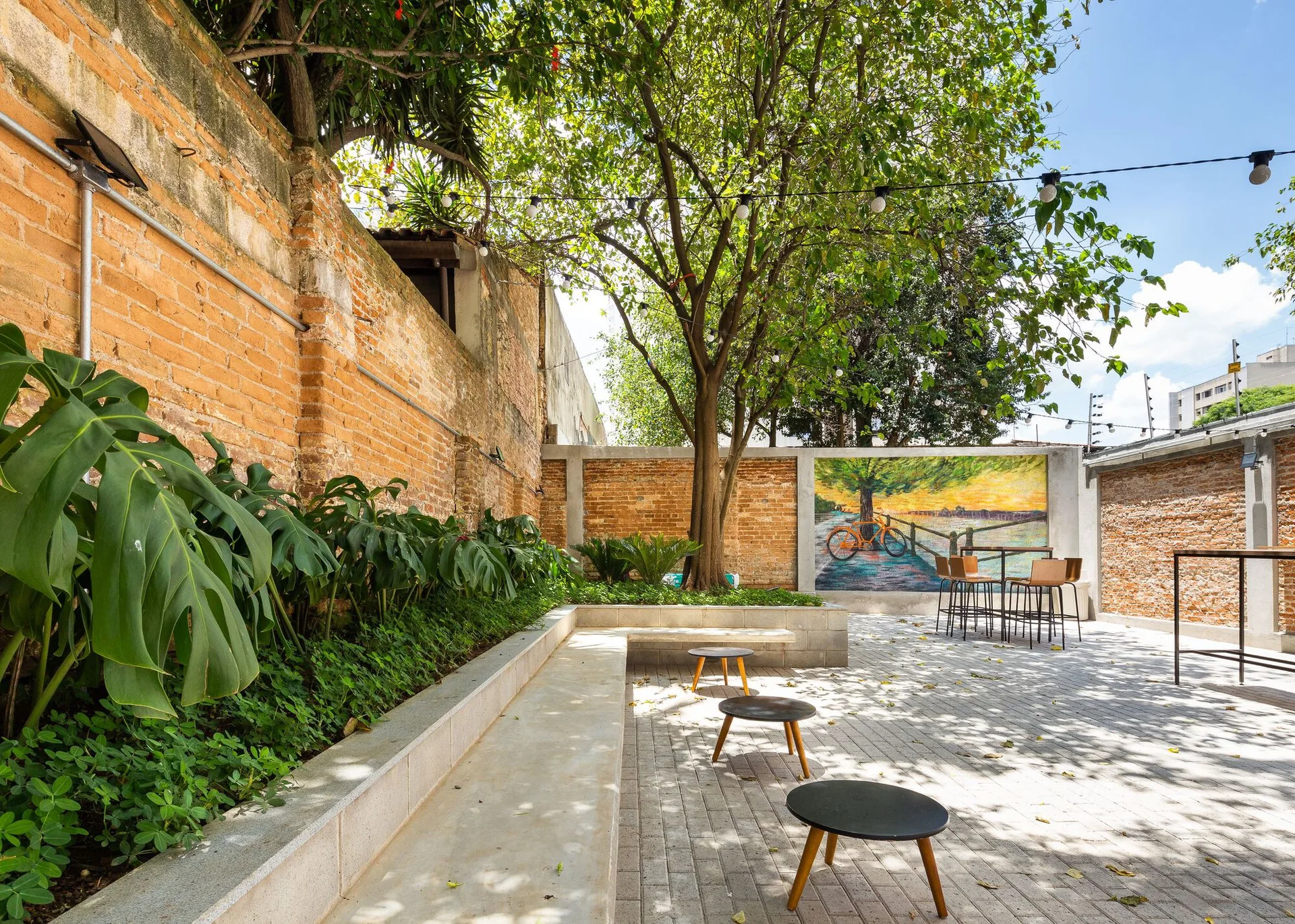 An outdoor brick patio featuring a built-in concrete bench planter, small round black tables, overhead string lights, and a colorful back wall mural.