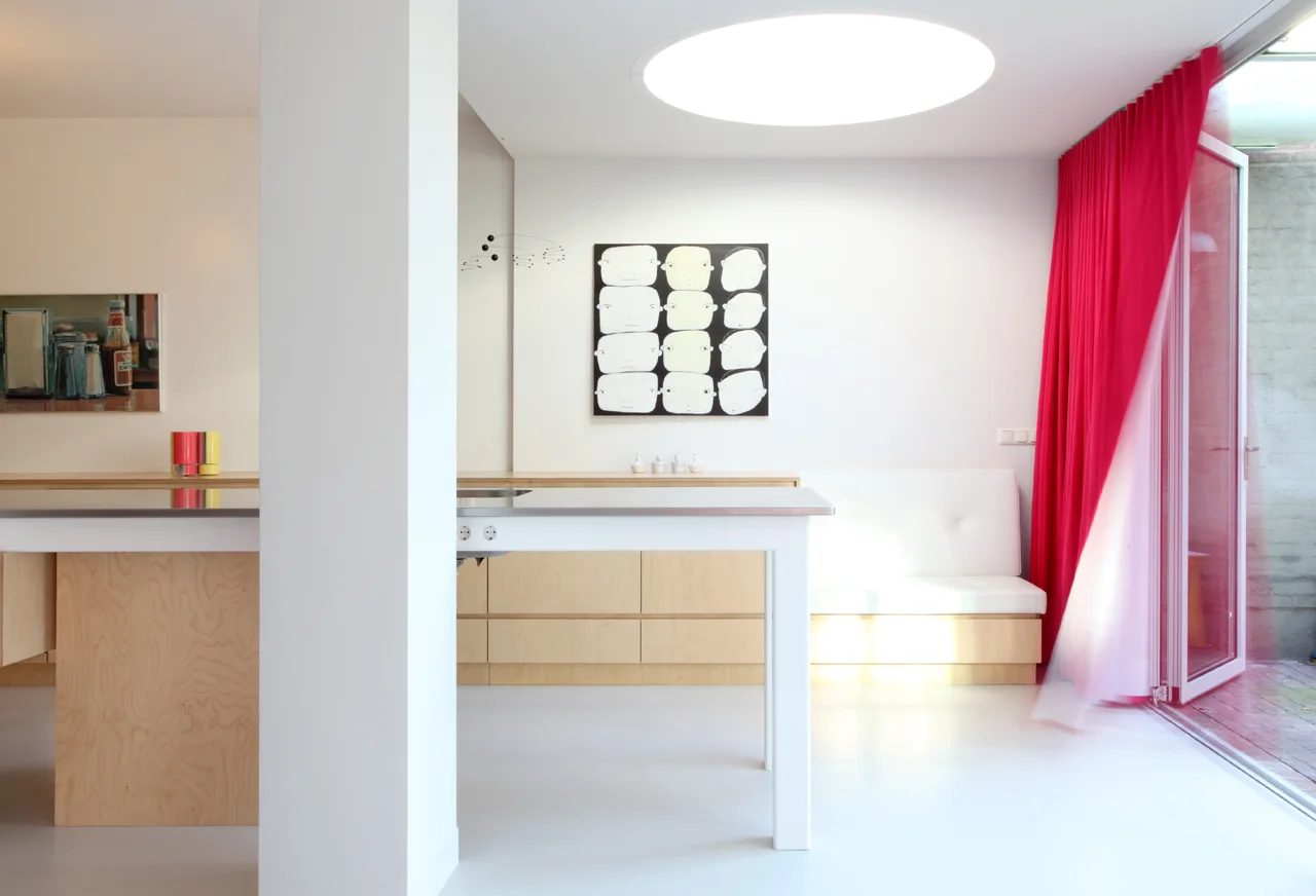 A minimalist kitchen with light plywood cabinets, a white banquette, a large circular skylight, and a vibrant magenta curtain next to a glass door.