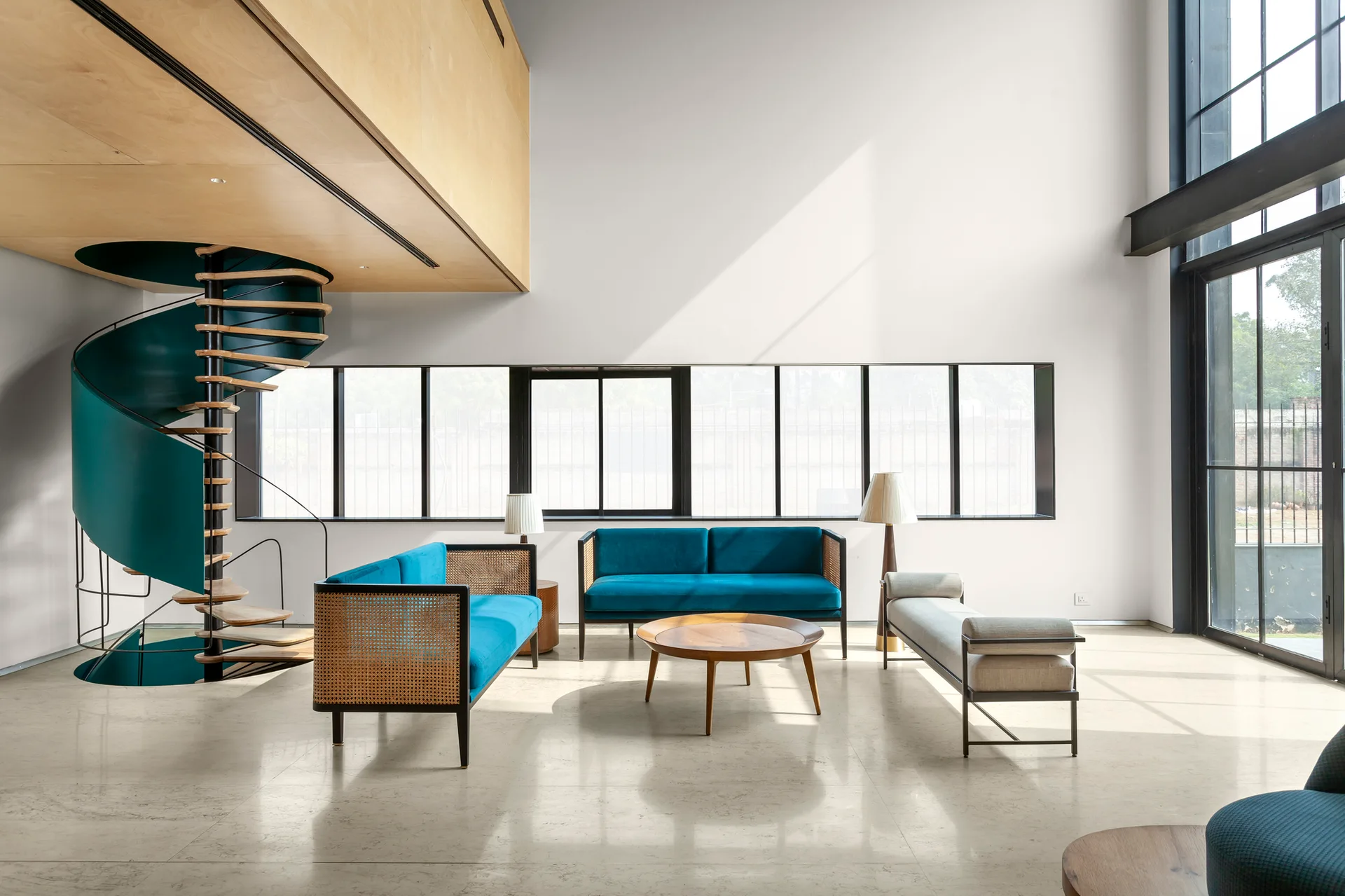 A spacious double-height living room featuring a teal spiral staircase, two blue cane-backed sofas, and a polished stone floor.