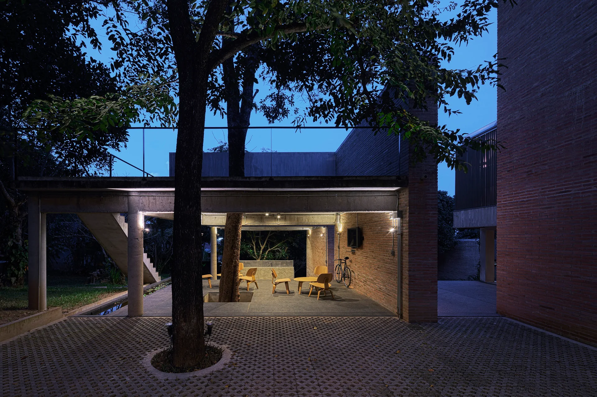 A covered concrete patio featuring a large tree growing through the structure, wood chairs, and an exposed brick wall at twilight.