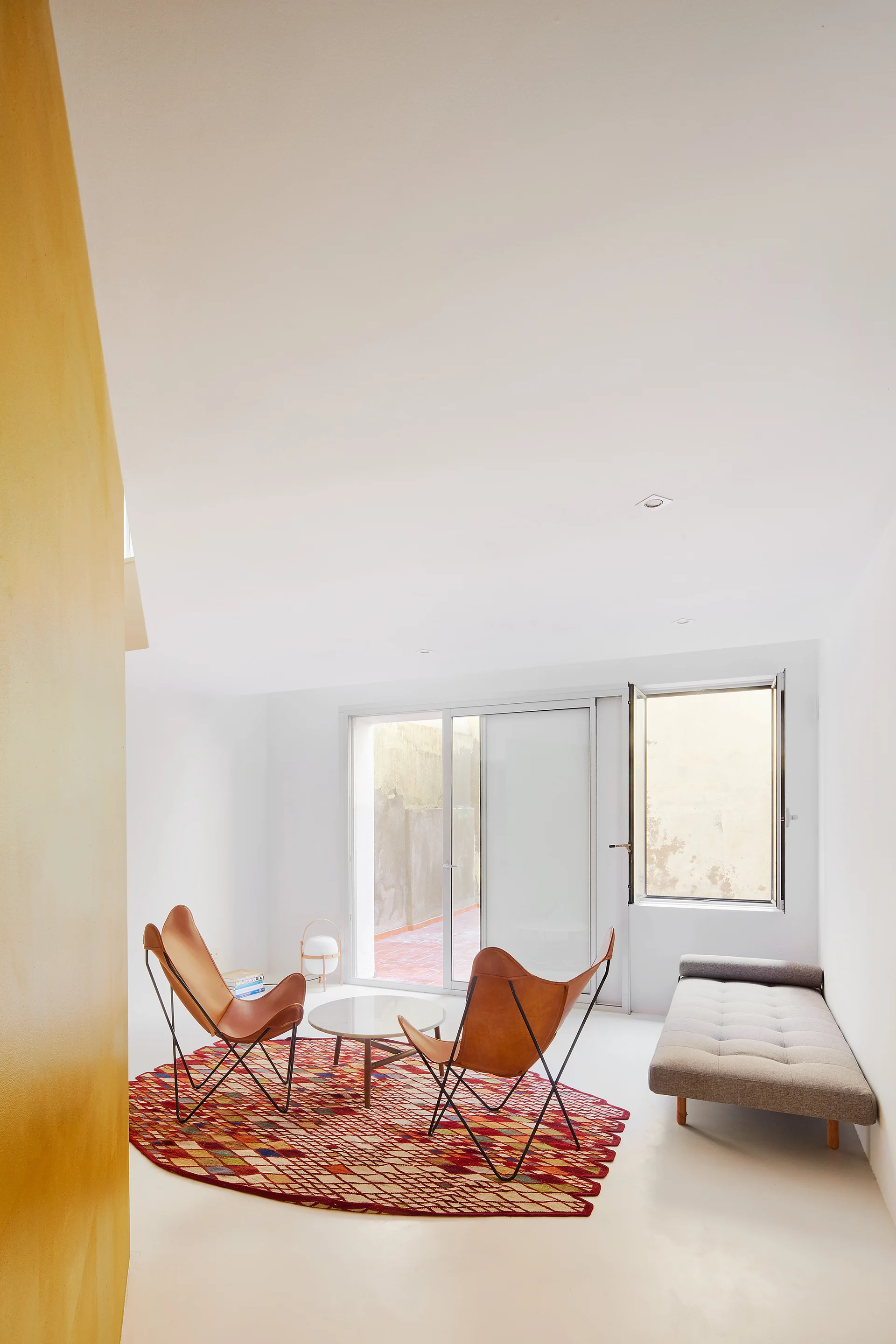 Two tan leather butterfly chairs sit on a red patterned rug next to a grey daybed in a minimalist white living room.