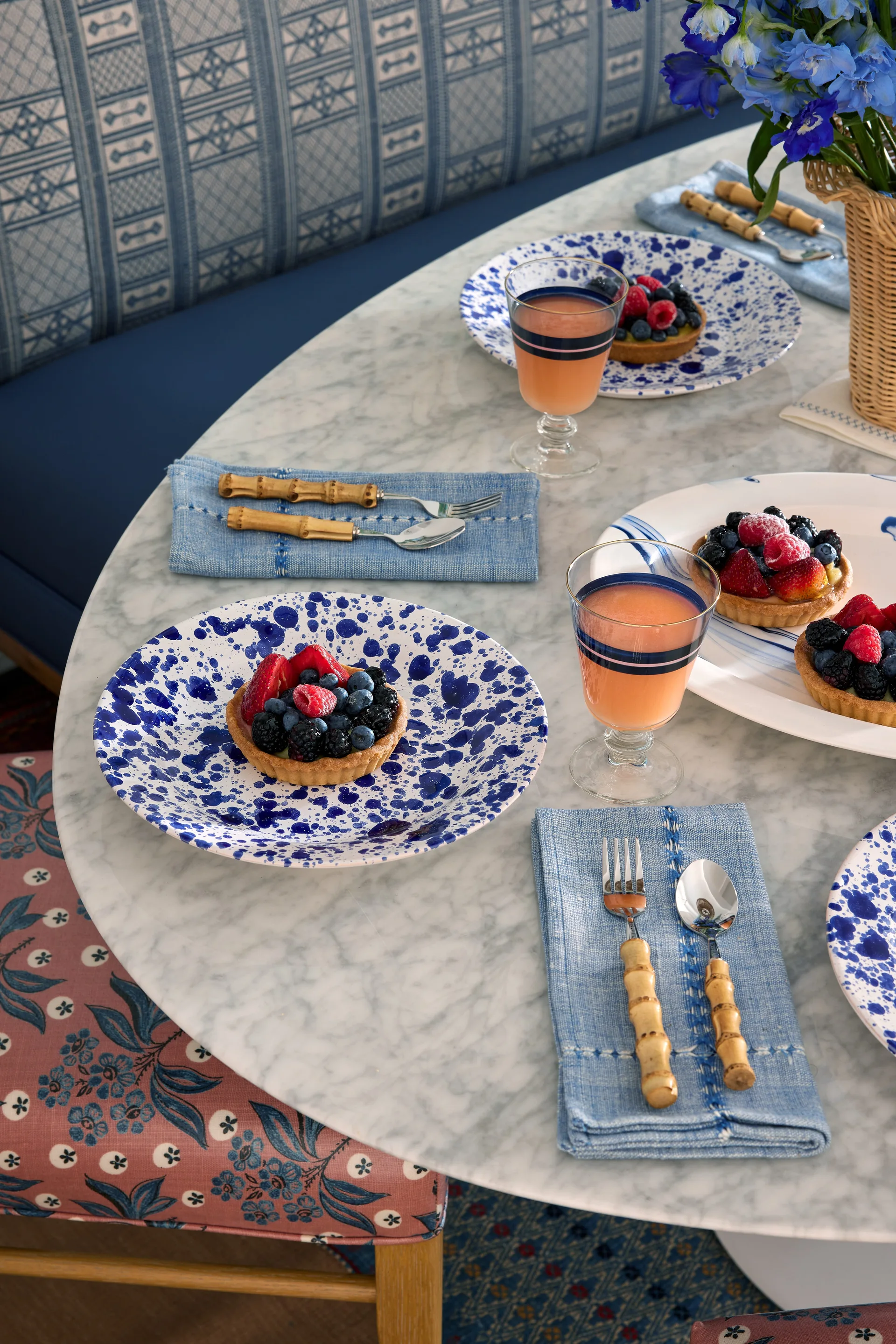 A round marble dining table set with blue and white speckled plates, bamboo-handled silverware, and berry tarts next to a blue patterned banquette.
