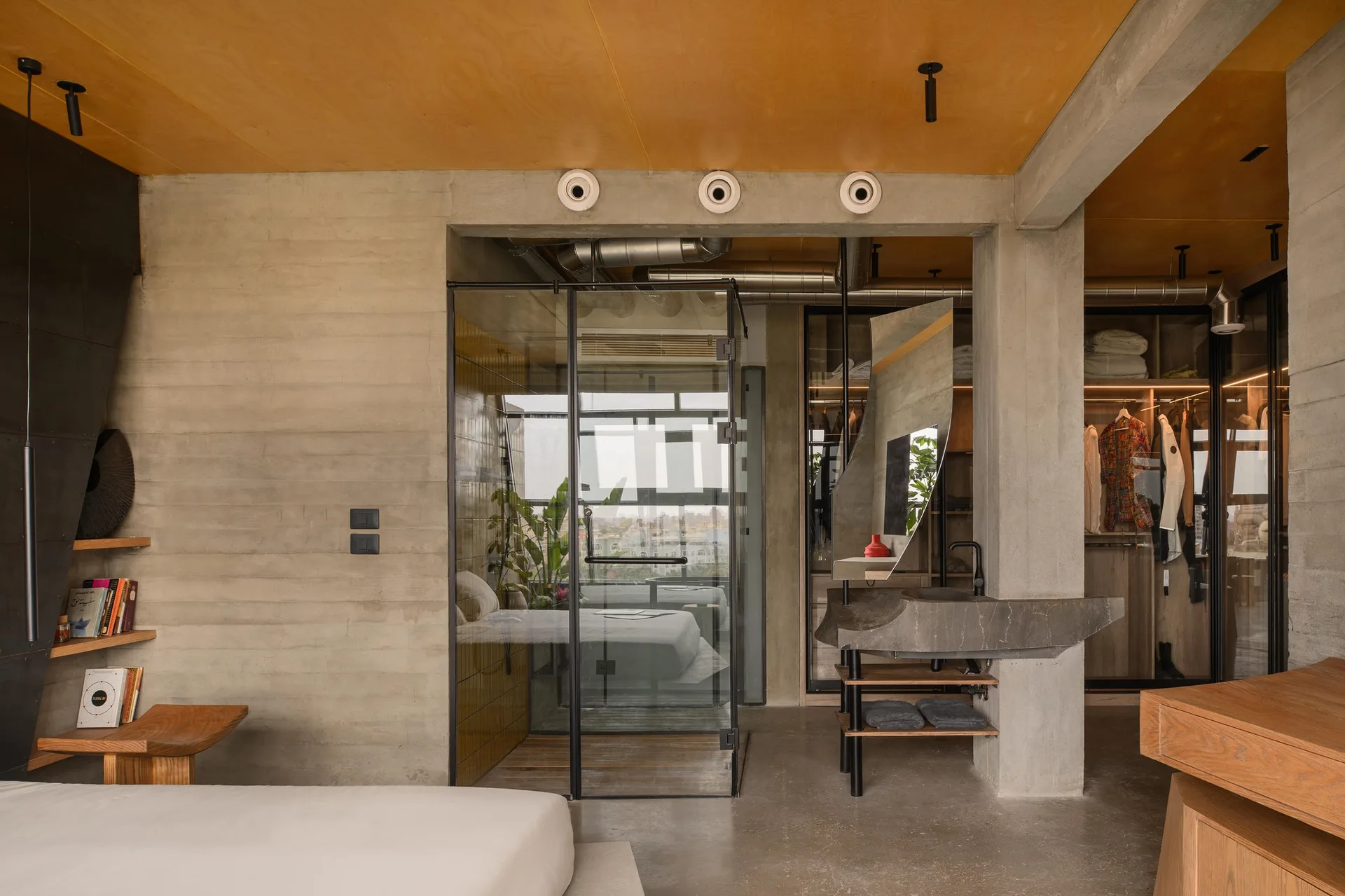 An industrial loft bedroom features board-formed concrete walls, a plywood ceiling, and a glass-enclosed bathroom alongside a concrete vanity.