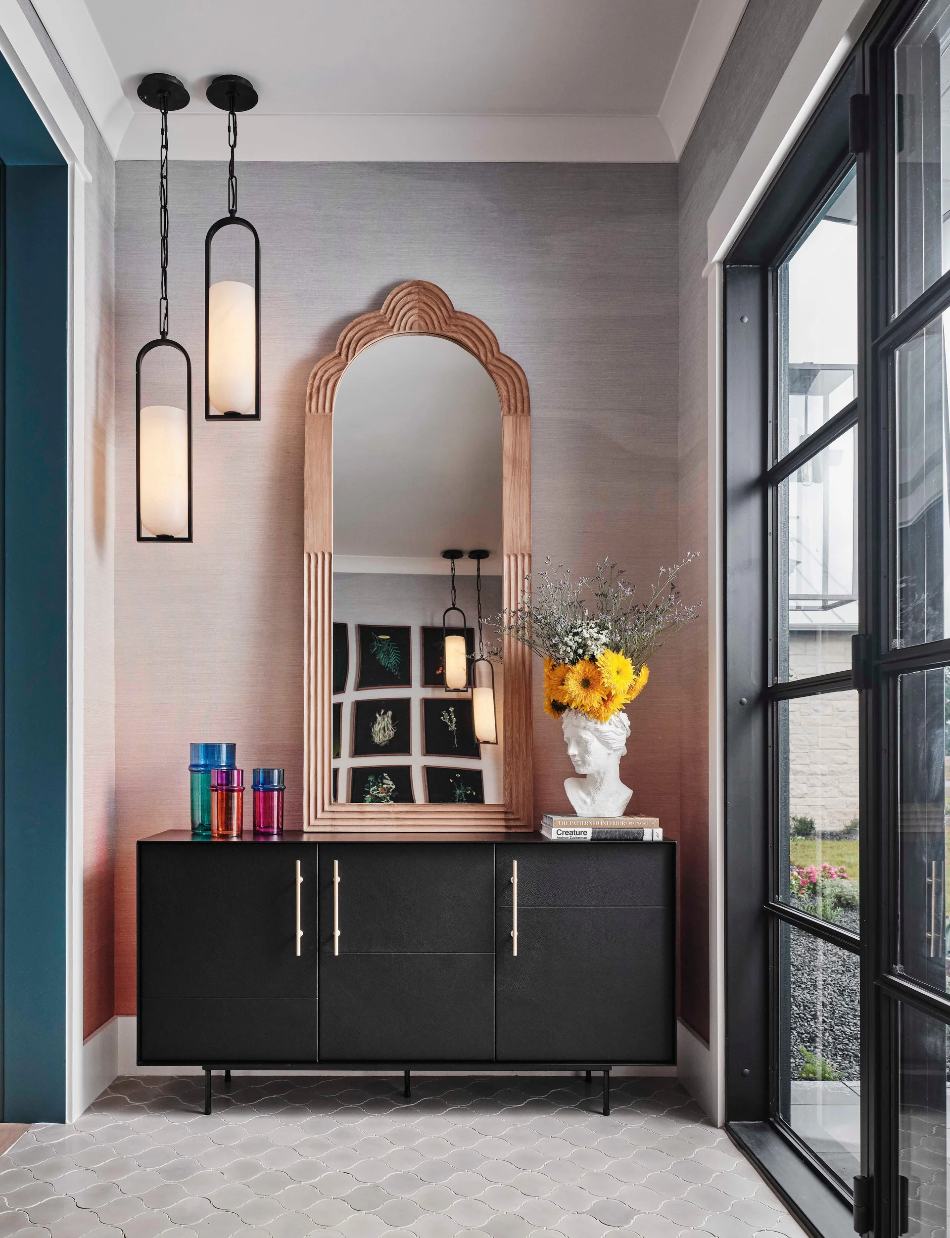 A modern entryway features a black console table under a tall arched wood mirror, set against a pink-and-grey gradient wall with scalloped tile flooring.