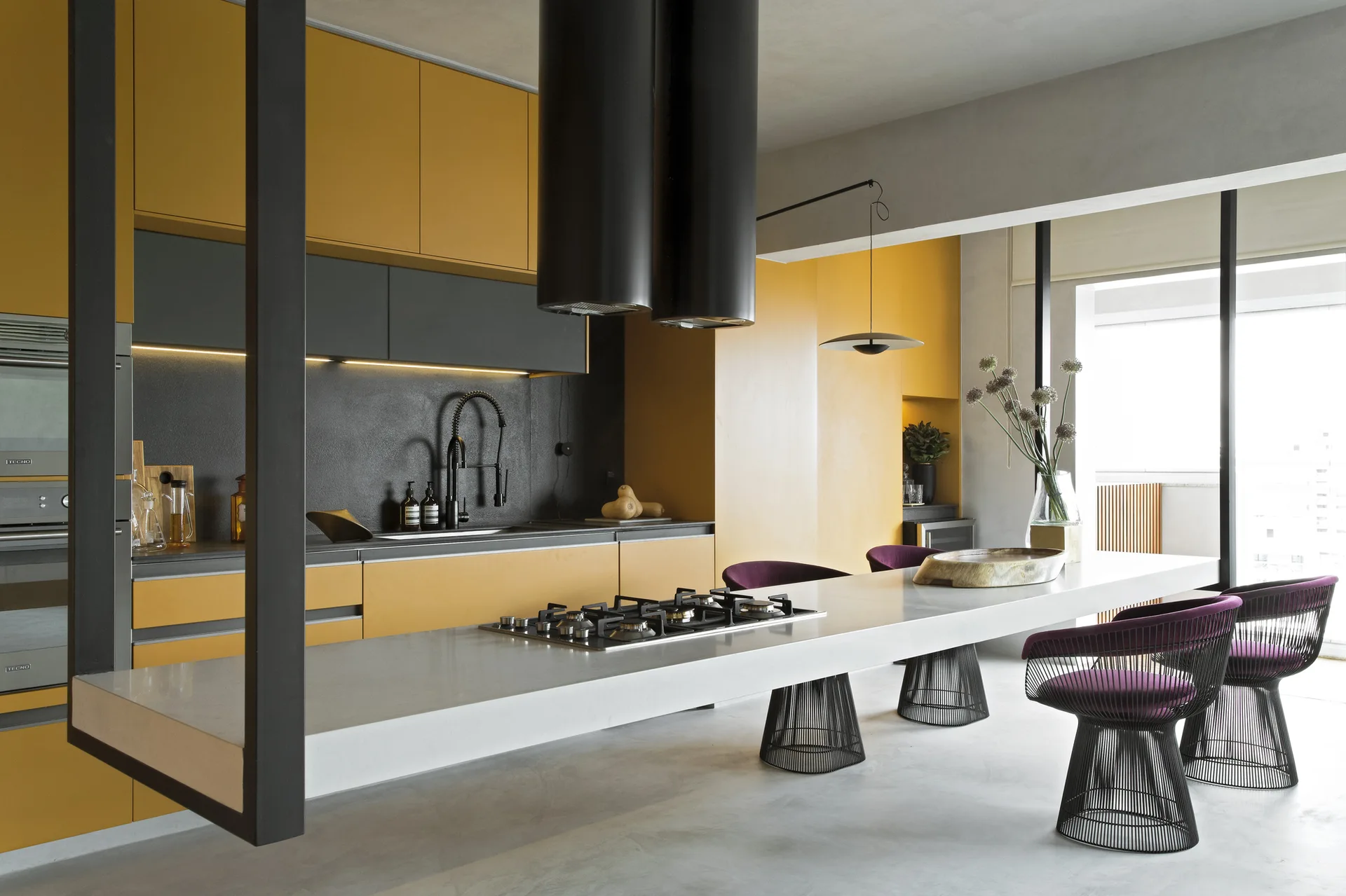 A modern kitchen featuring matte mustard yellow cabinets, a long white countertop island with integrated stovetop, and purple wire-frame bar stools.