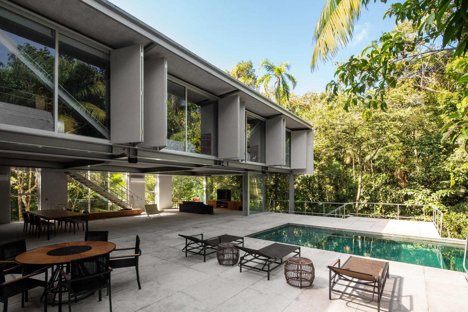 A modern concrete patio features a rectangular swimming pool, metal sun loungers, and dining tables beneath a two-story steel and glass building.