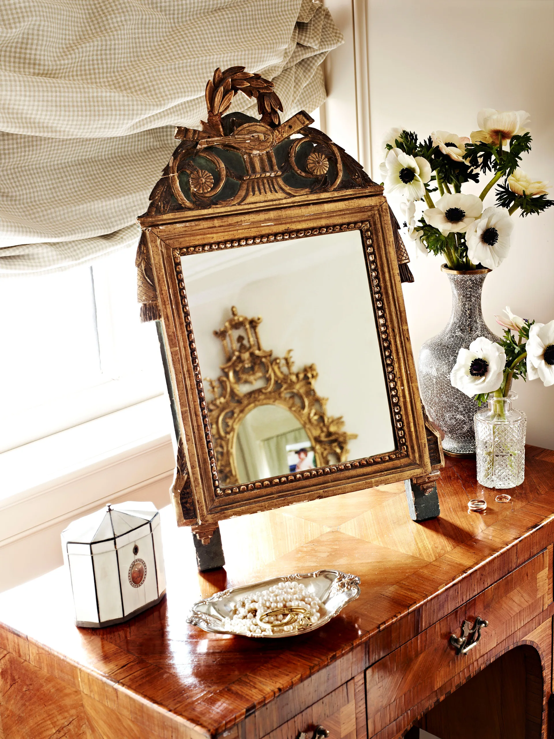 An ornate gilt table mirror sits on a polished wood vanity alongside vases of white anemone flowers, a silver tray of pearls, and a lidded box.