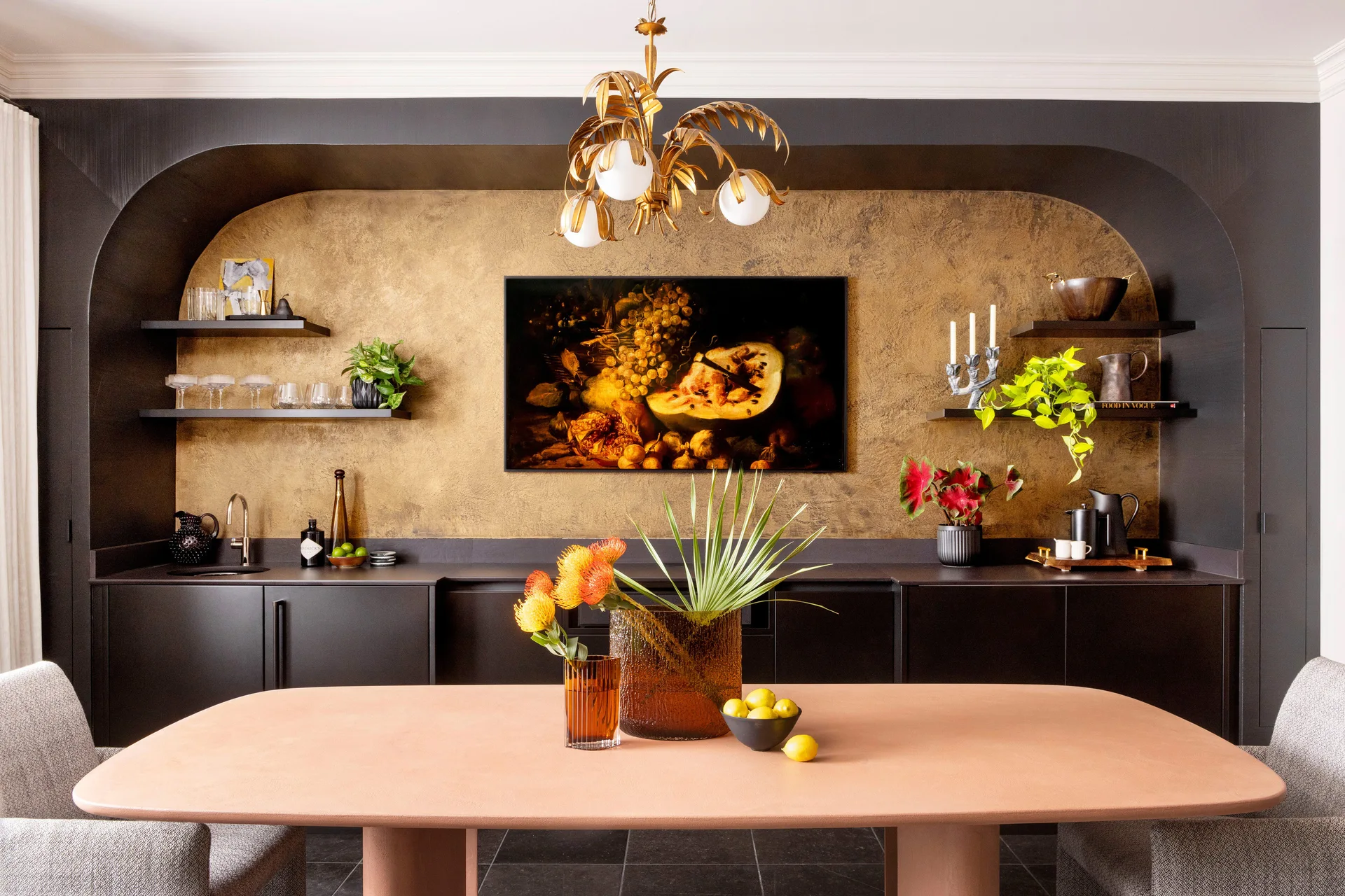 A peach-colored dining table sits in front of a black built-in credenza featuring a recessed gold arch, floating shelves, and still life artwork.