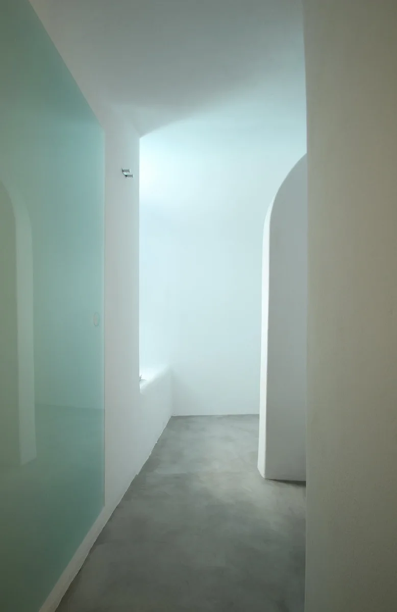 A minimalist bathroom entryway features smooth white plaster walls, a curved archway, a light blue glass partition, and matte grey concrete flooring.