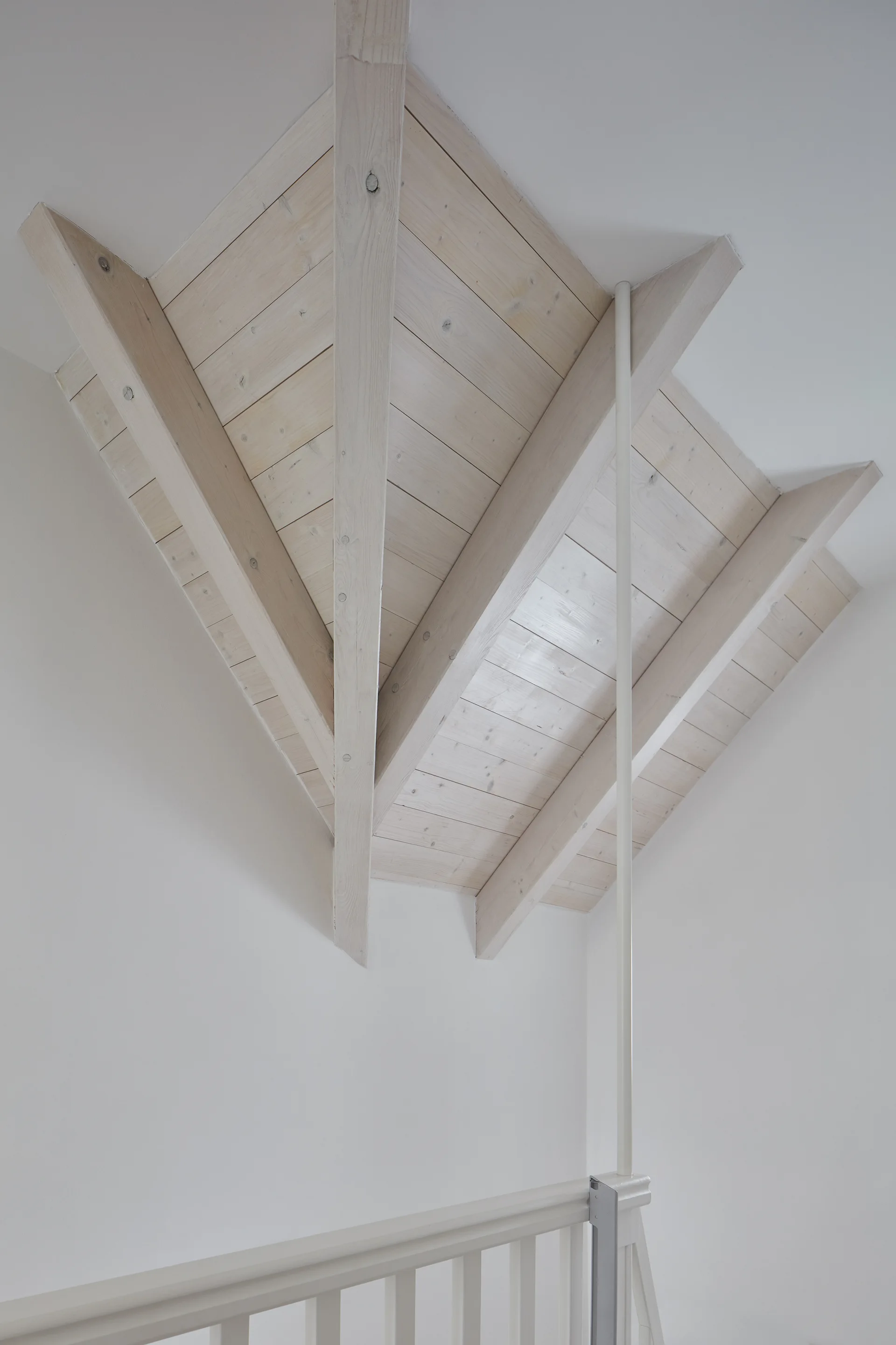 A low-angle view of a sloped whitewashed wood plank ceiling with exposed rafters above a white painted staircase railing.