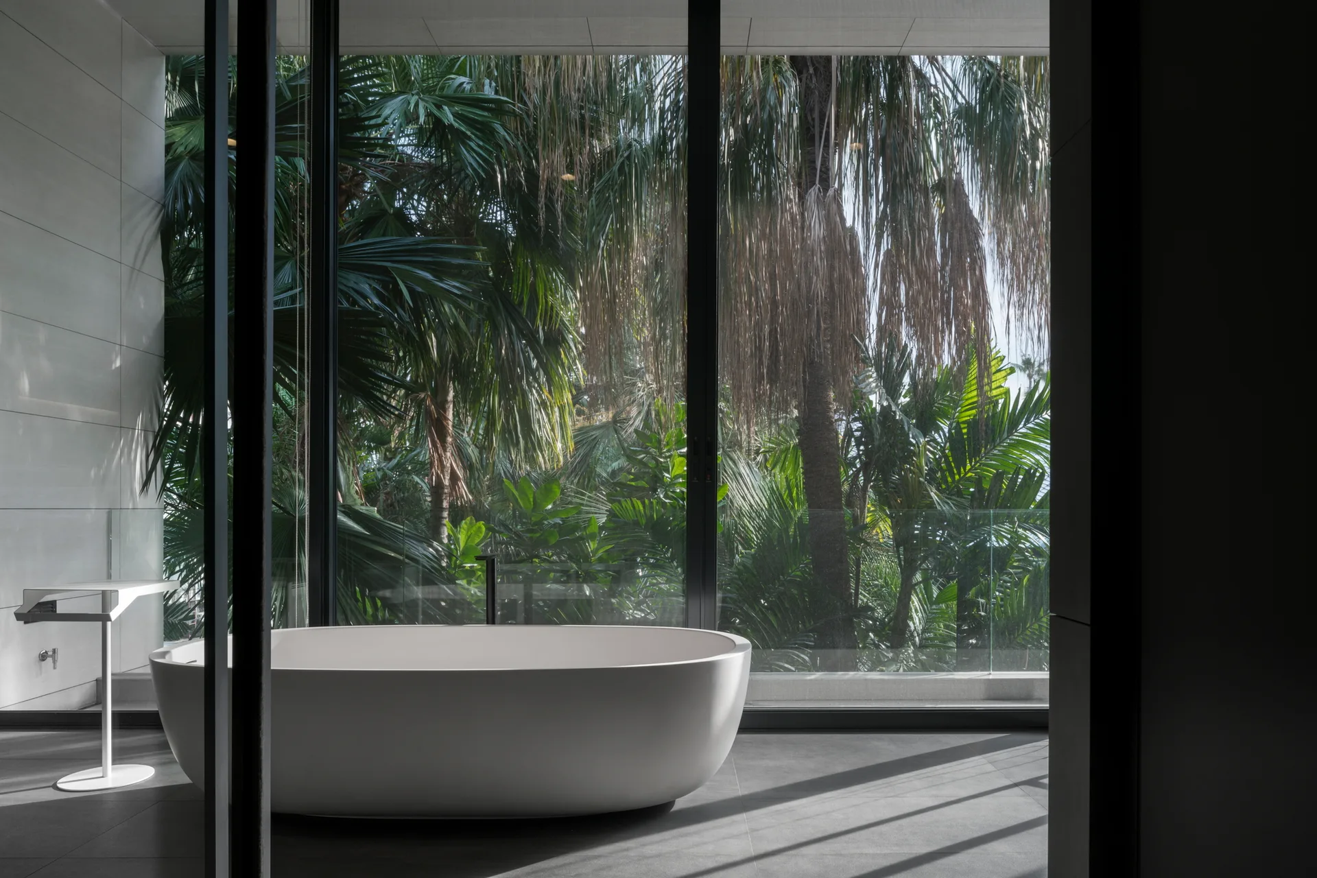 A white freestanding oval bathtub sits on dark gray tiled floors in front of a floor-to-ceiling window overlooking lush tropical green palm trees.