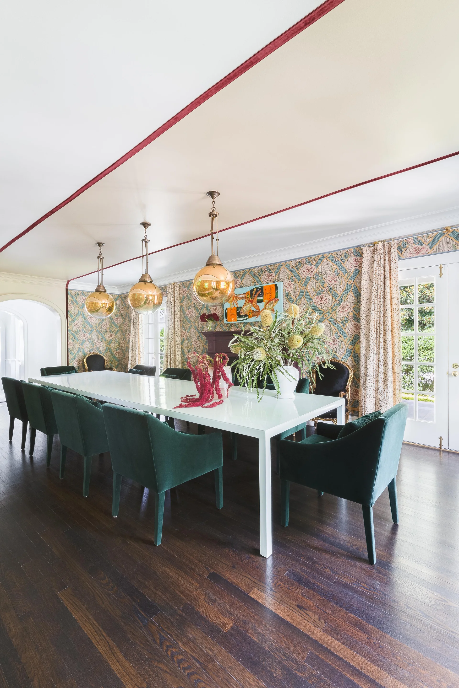 A formal dining room features green velvet armchairs around a long white table beneath three brass globe pendant lights and floral patterned wallpaper.