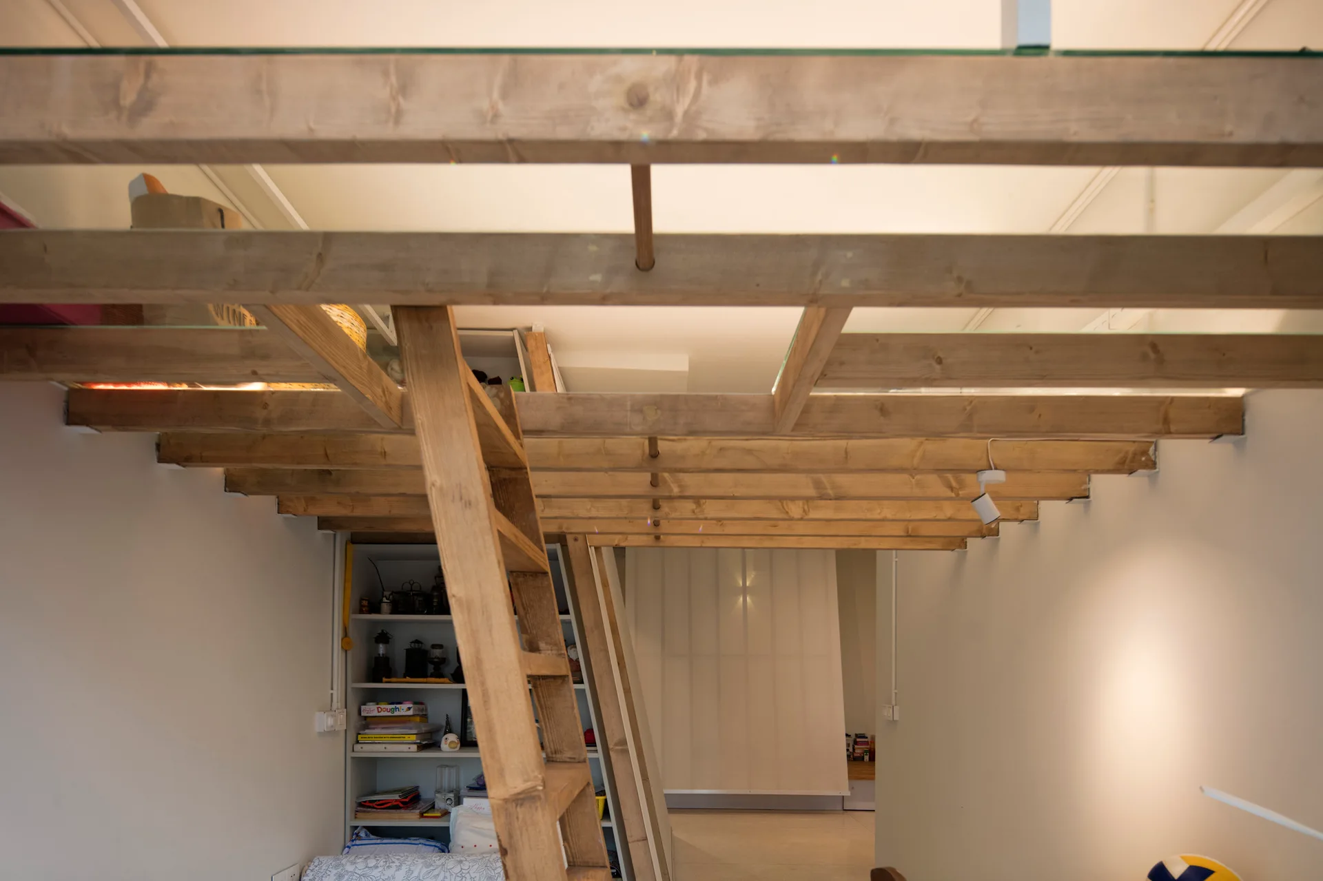 A low-angle view of a wooden mezzanine loft featuring glass floor inserts, exposed timber joists, and a natural wood ladder leading up from a white room.