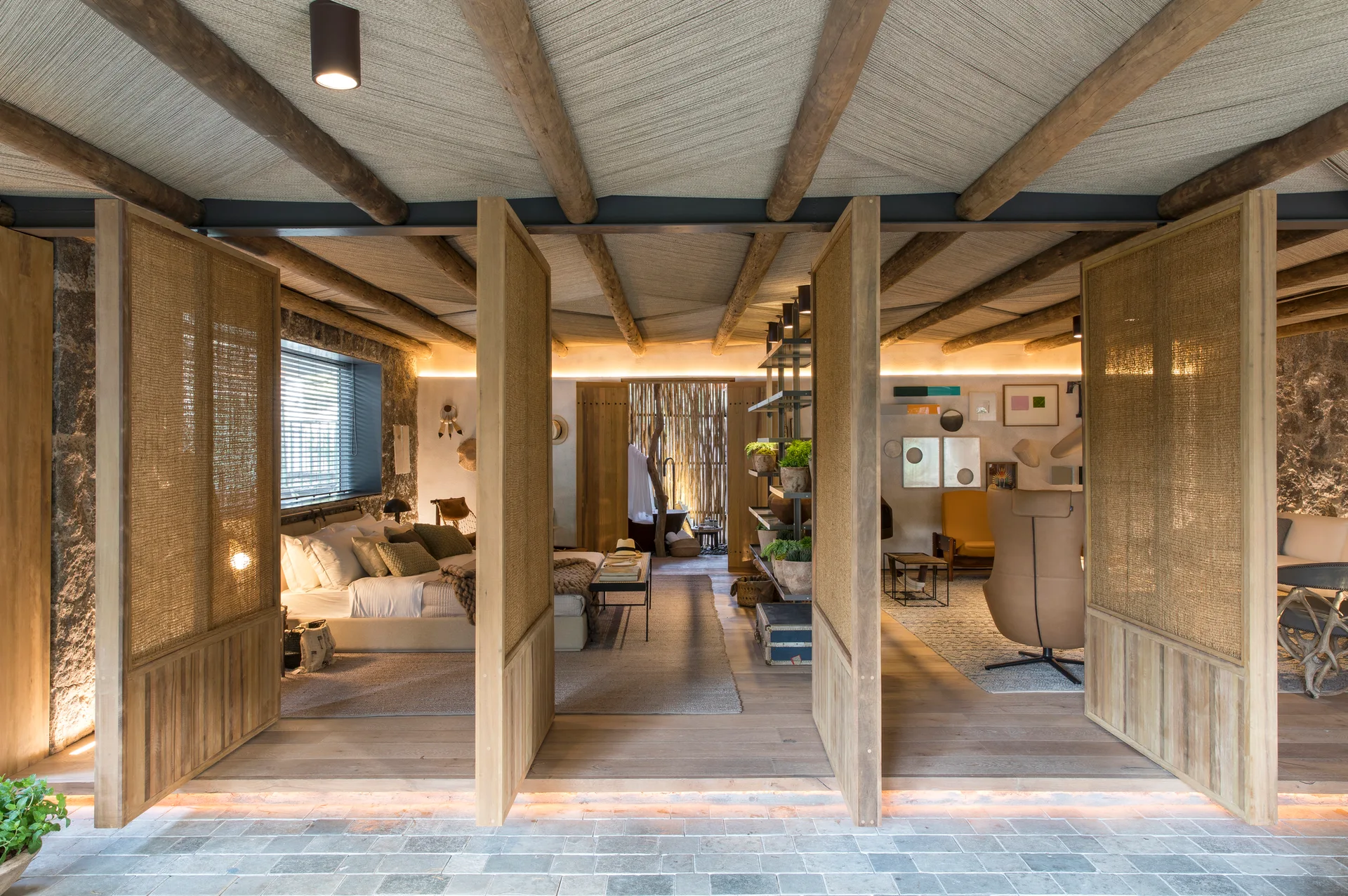 Large wooden screen dividers open to reveal a rustic bedroom and lounge area with exposed log ceiling beams, stone walls, and warm lighting.