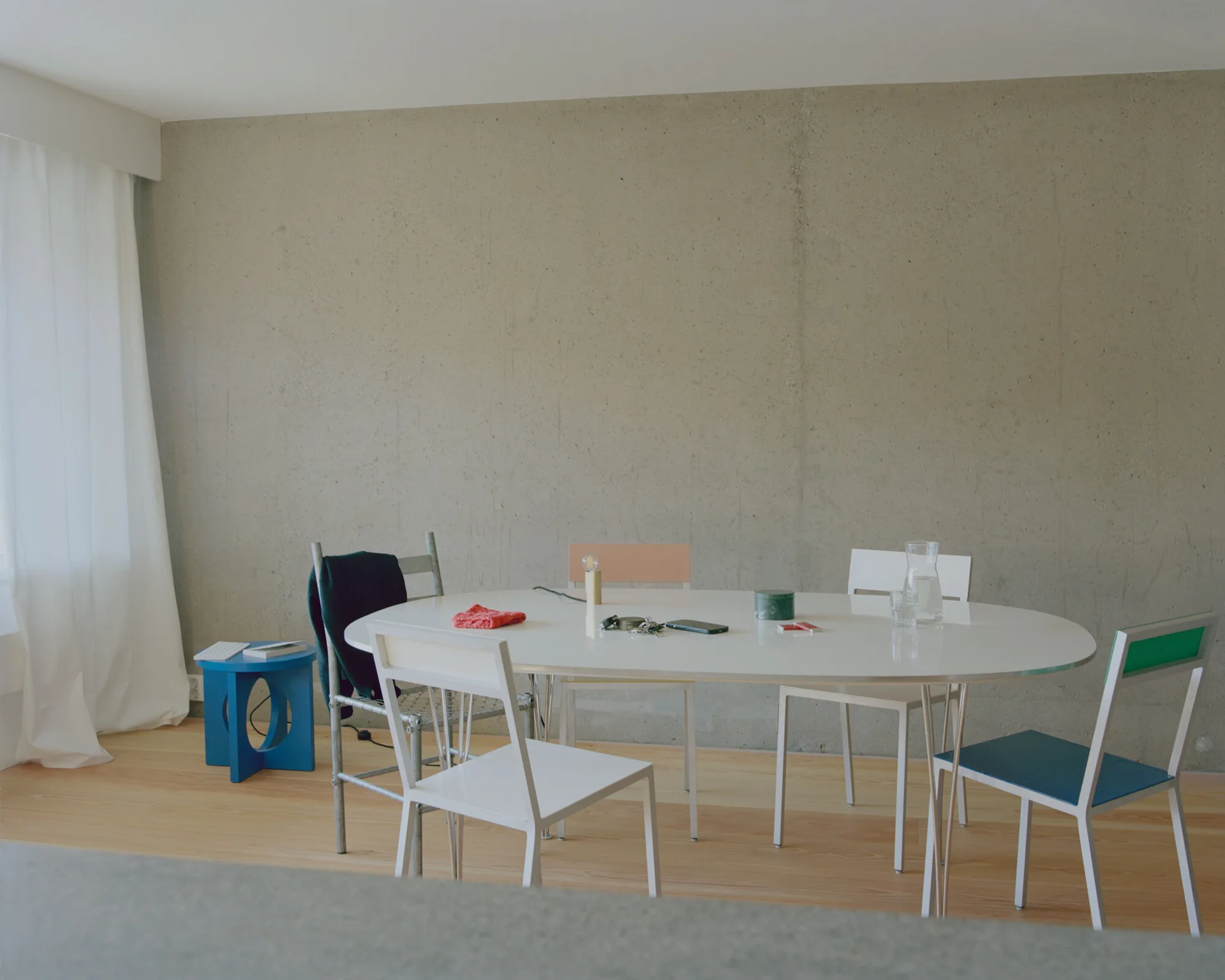 A minimalist dining room features an oval white table and mismatched chairs set against a tall, raw grey concrete wall with light wood floors.