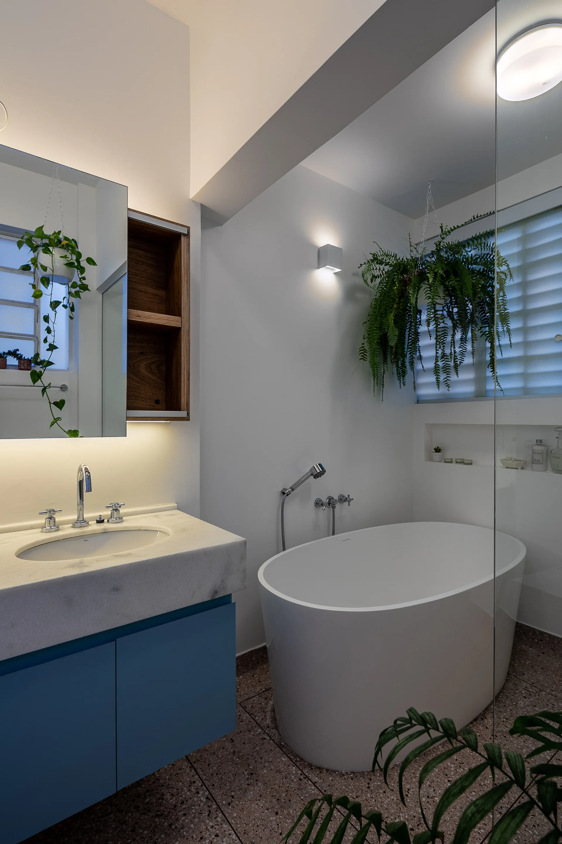 A modern bathroom features a white freestanding bathtub, a blue vanity with a stone countertop, speckled flooring, and lush hanging plants.