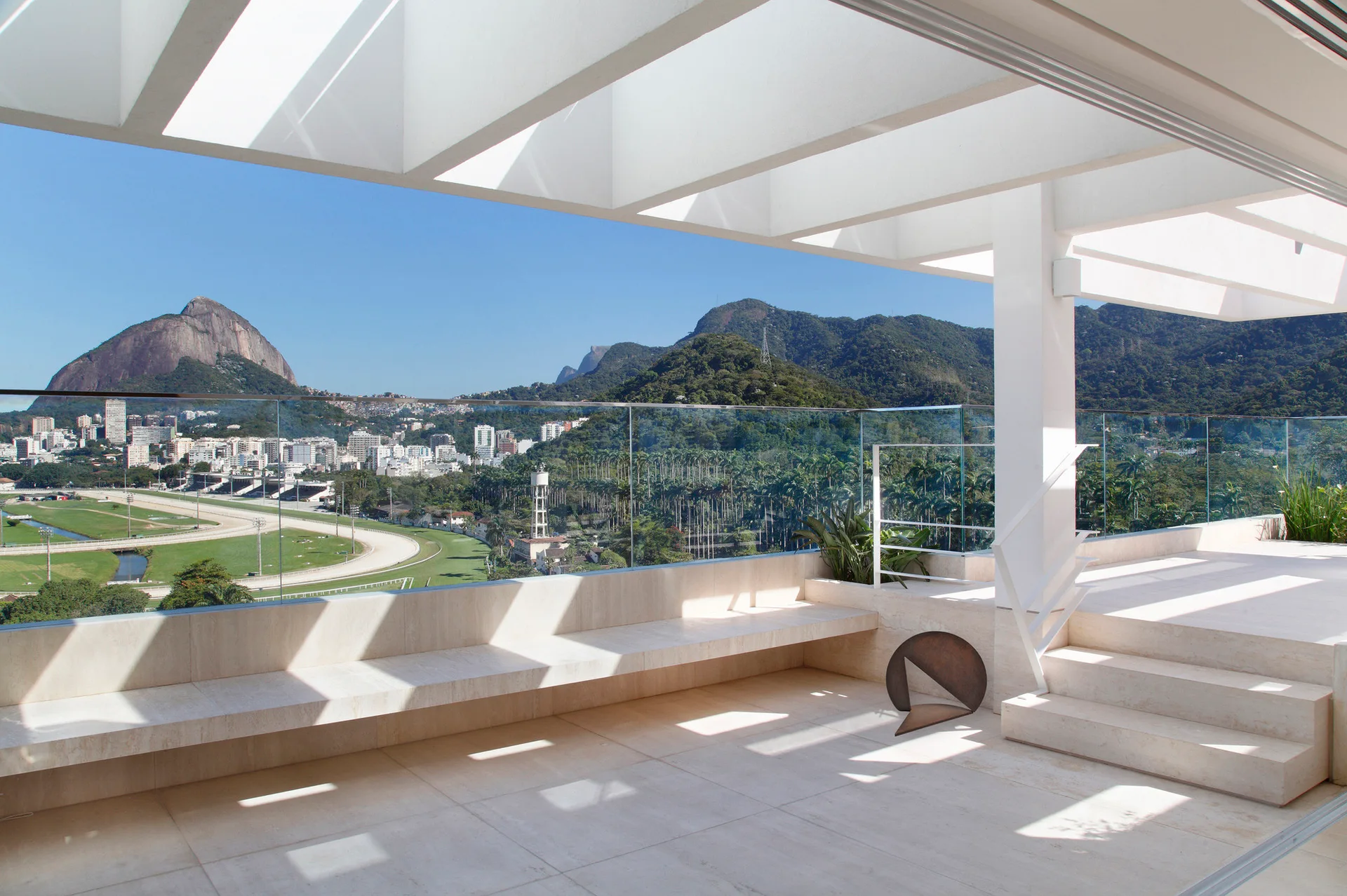 A beige stone terrace features an integrated bench, white slatted pergola, glass railing, and steps overlooking a city and mountain landscape.