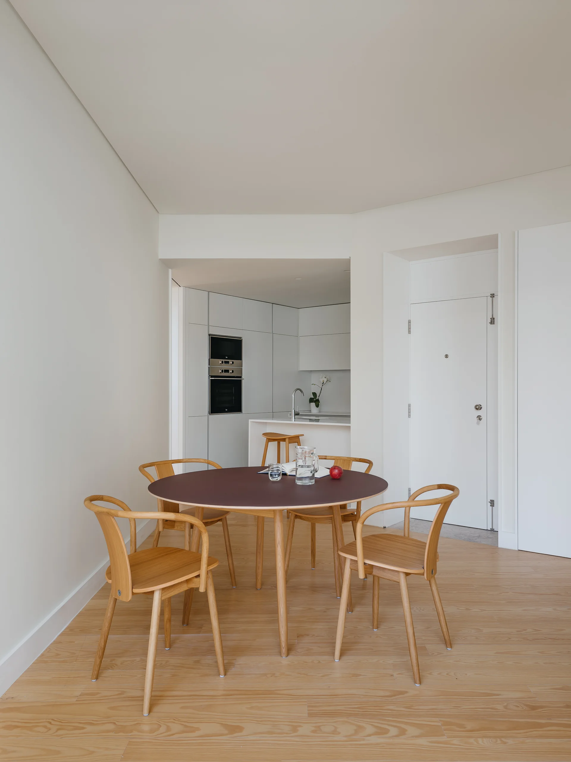A round dining table with a dark top and four light wood chairs sits on wood flooring in a bright dining area with a kitchen in the background.