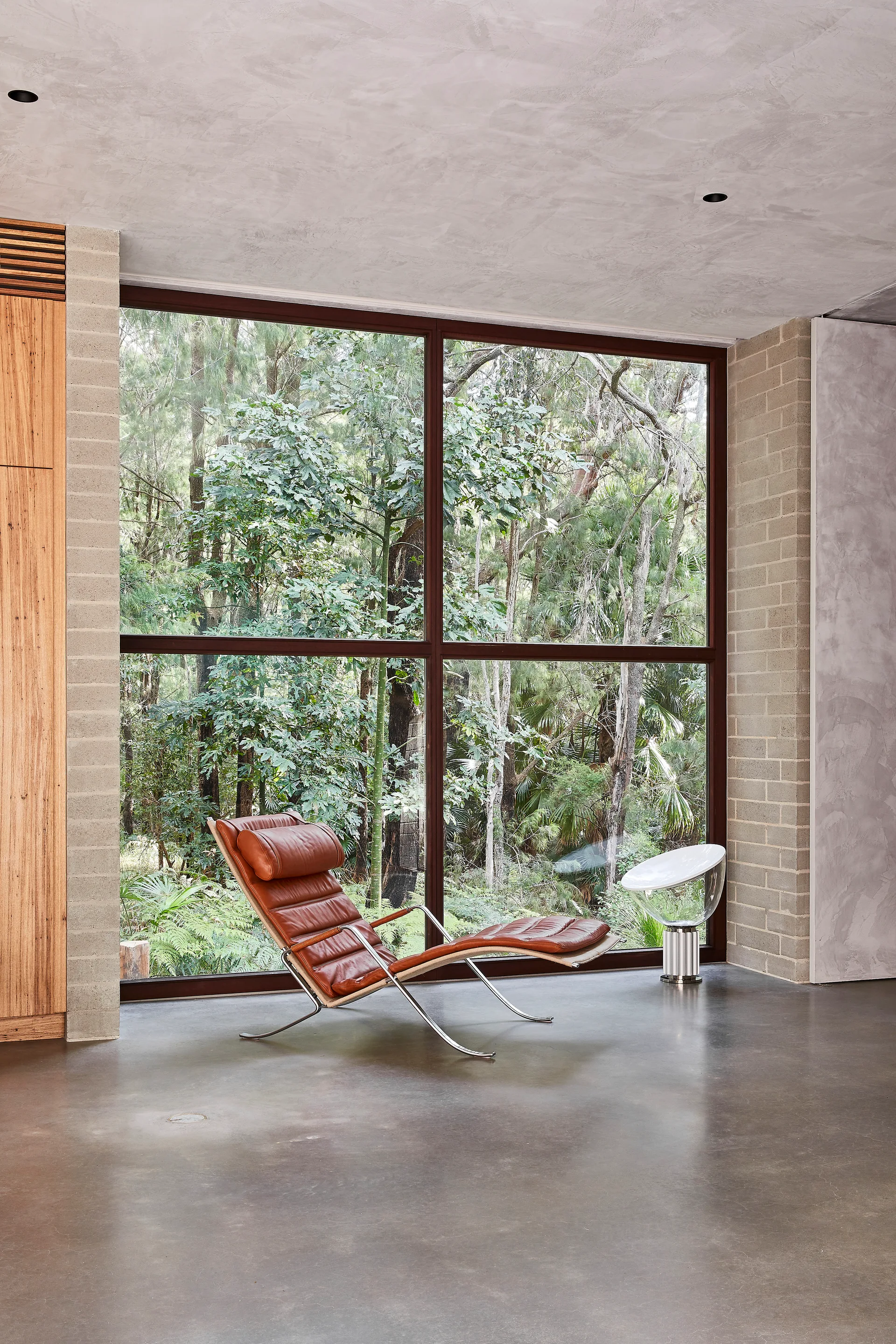 A brown leather chaise longue sits on a polished concrete floor near a large window showcasing a lush forest.