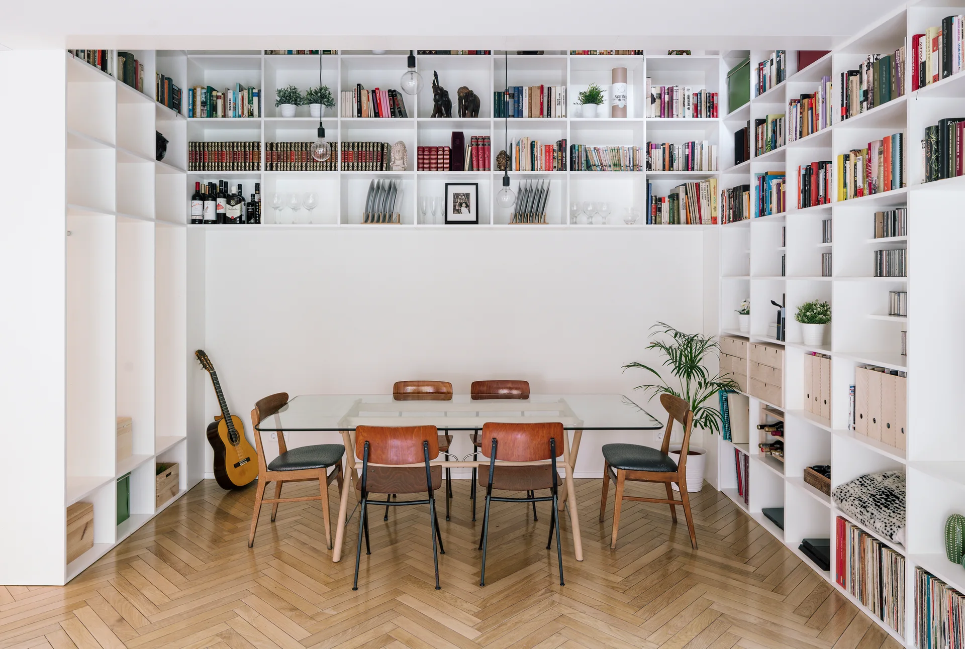 A dining area features a glass-top table with wooden chairs, set against a massive white built-in bookshelf on herringbone hardwood floors.